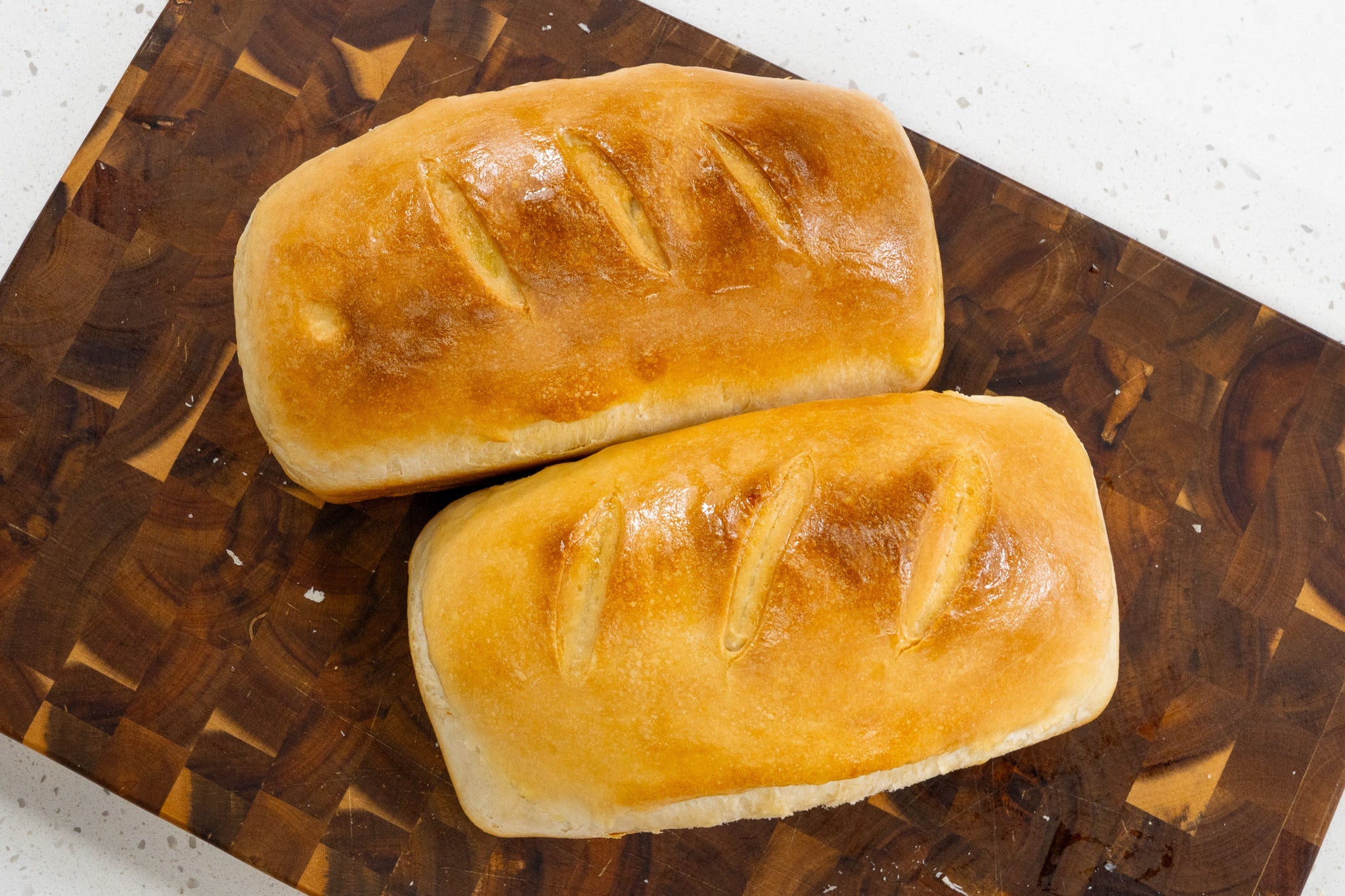 Two golden-brown loaves of sourdough sandwich bread with three decorative slashes each, cooling on a dark wooden cutting board.