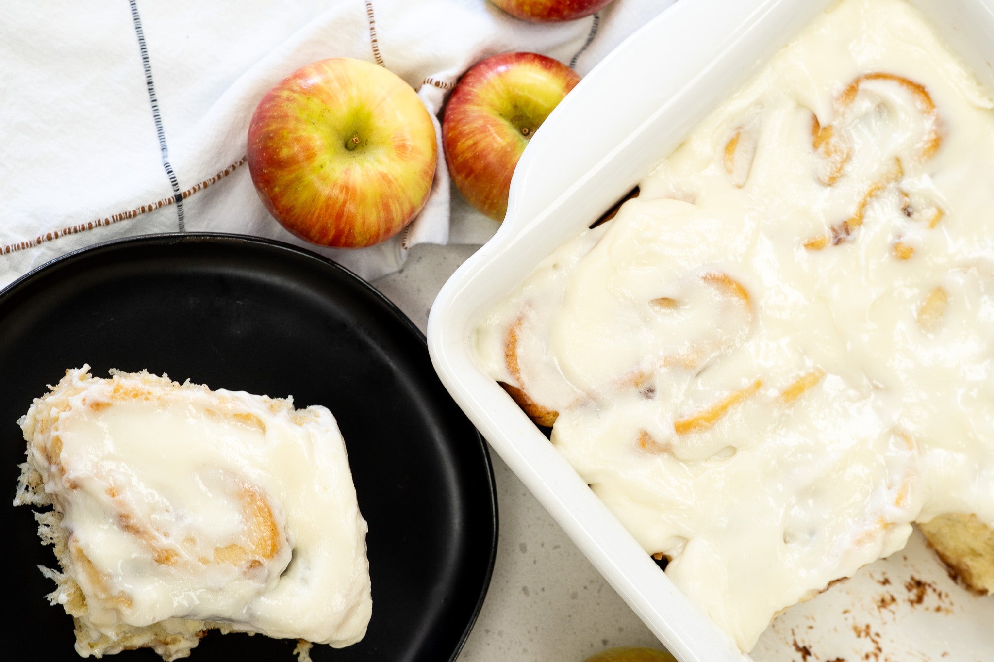 Freshly baked apple pie cinnamon rolls in a white baking dish, topped with cream cheese frosting, with one roll served on a black plate and fresh apples placed alongside on a white cloth.