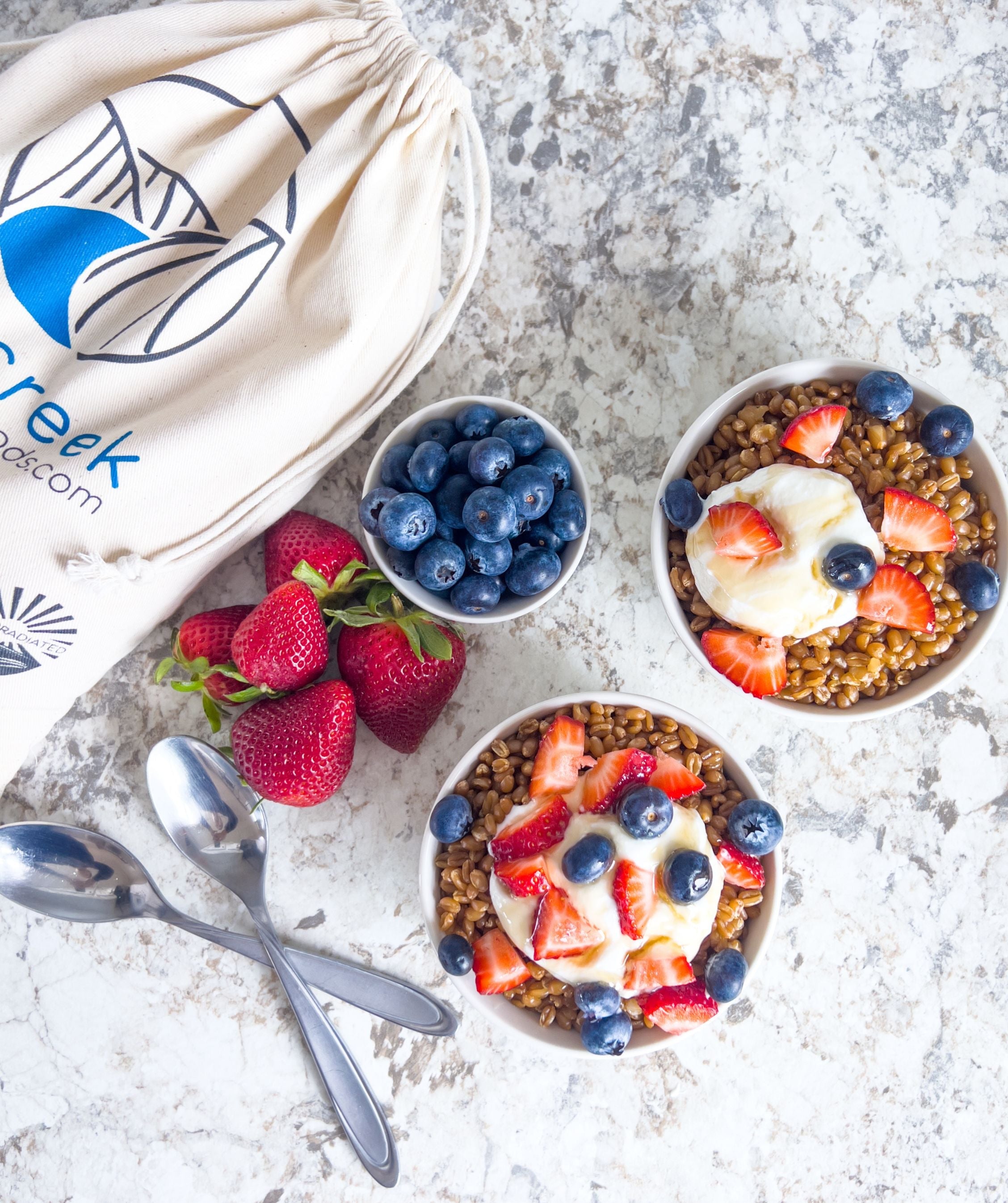 Two cups of grains with whipped cream, berries, and honey on a grey counter next to strawberries, blueberries, spoons, and a bag of grains