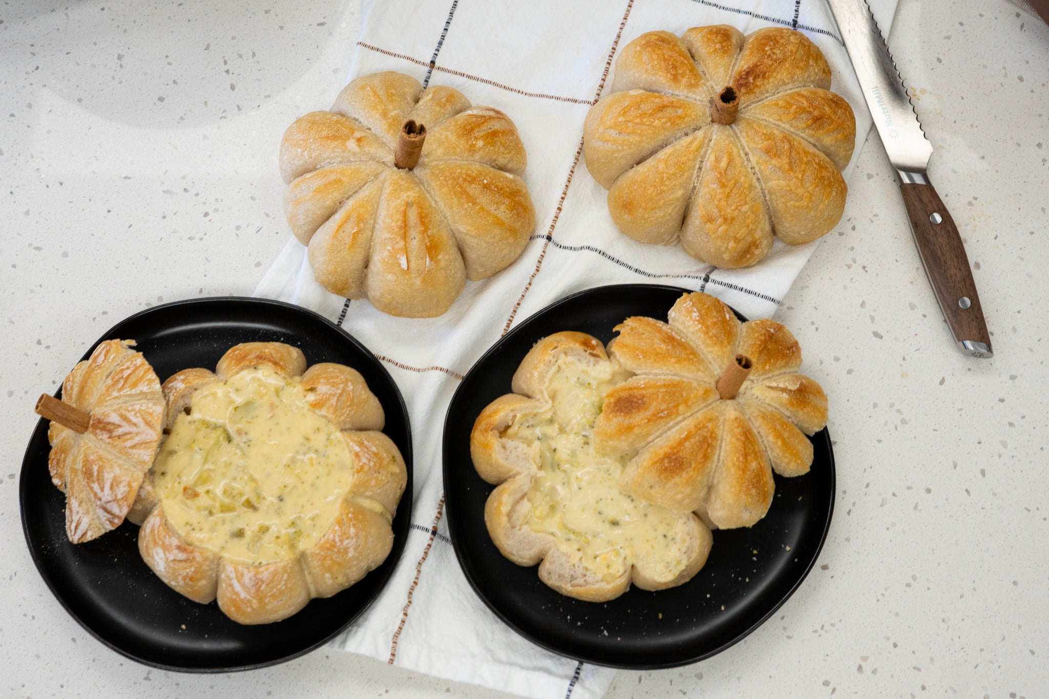 Two pumpkin-shaped bread loaves stuffed with soup, sitting on black plates below two pumpkin-shaped bread lids next to a bread knife, all sitting on a white towel on a white speckled counter