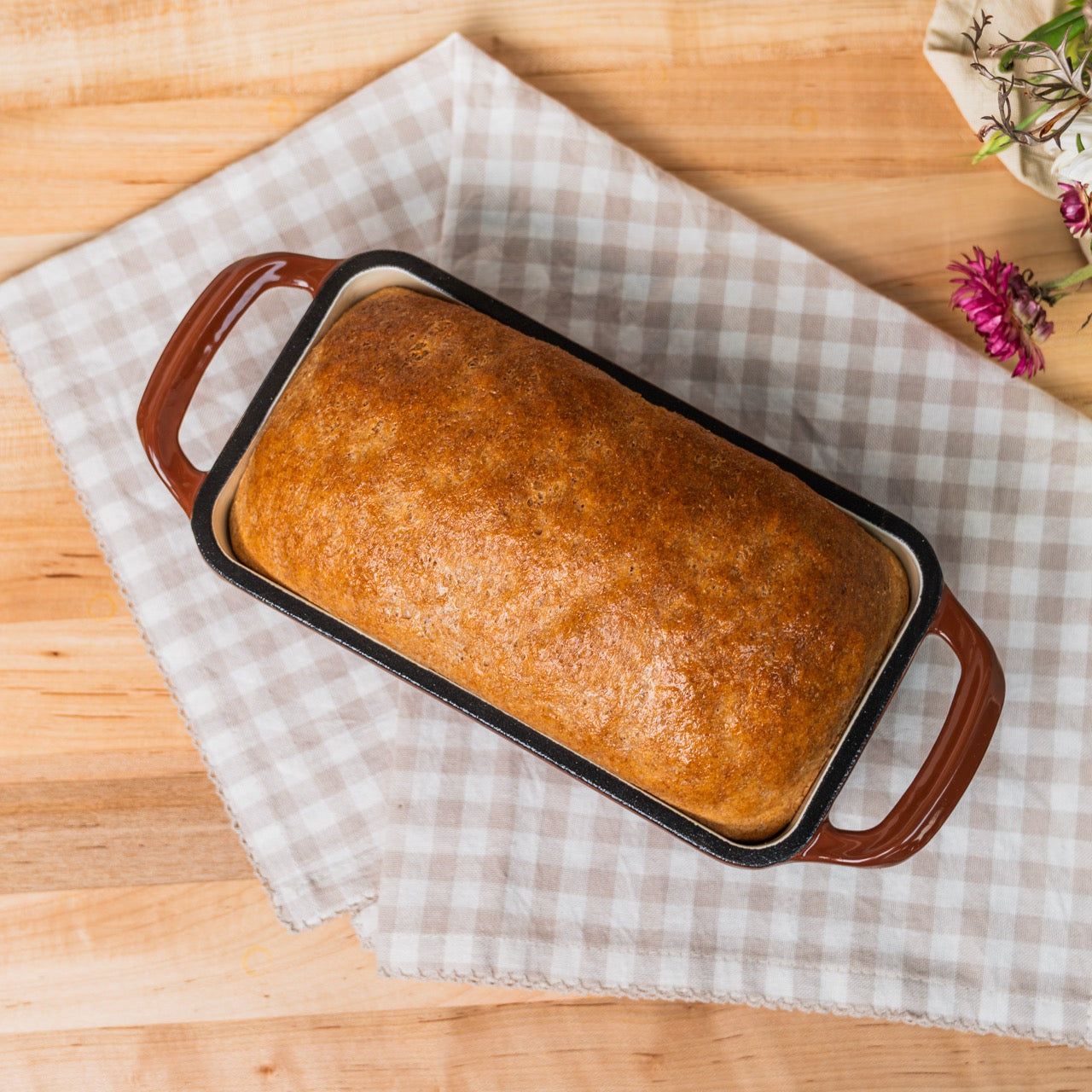 Loaf of bread in a brown baking dish on a checkered cloth with a wooden surface.