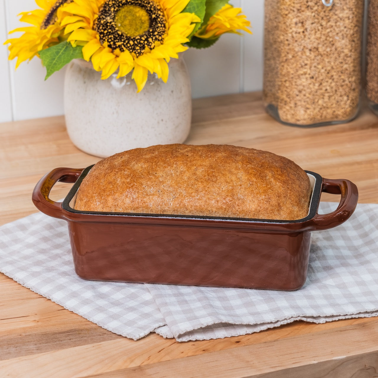 Loaf of bread in a brown ceramic dish on a wooden surface with sunflowers in the background.