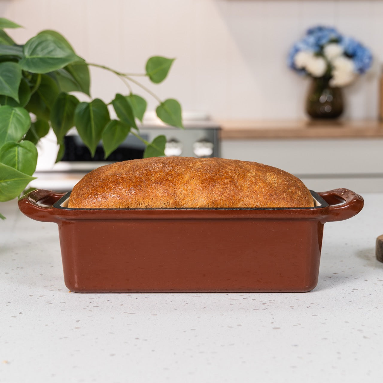 Loaf of bread in a brown ceramic loaf pan on a kitchen counter with a plant and flowers in the background.