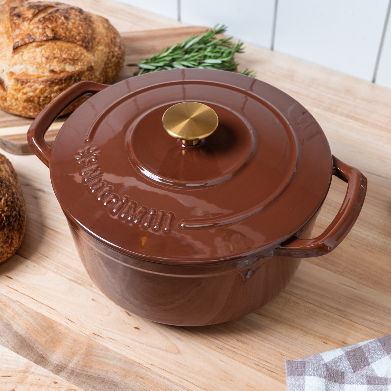 Brown enameled cast iron pot with a gold handle on a wooden surface with bread and herbs.