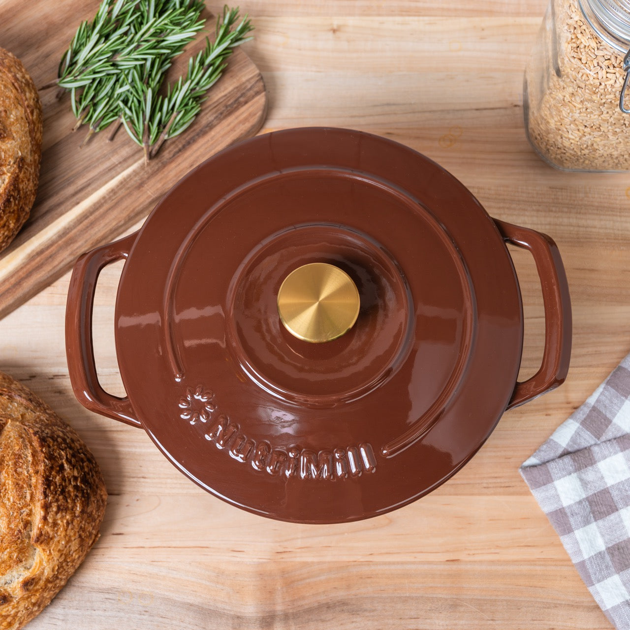 Brown ceramic pot with gold knob on a wooden surface with bread and herbs.