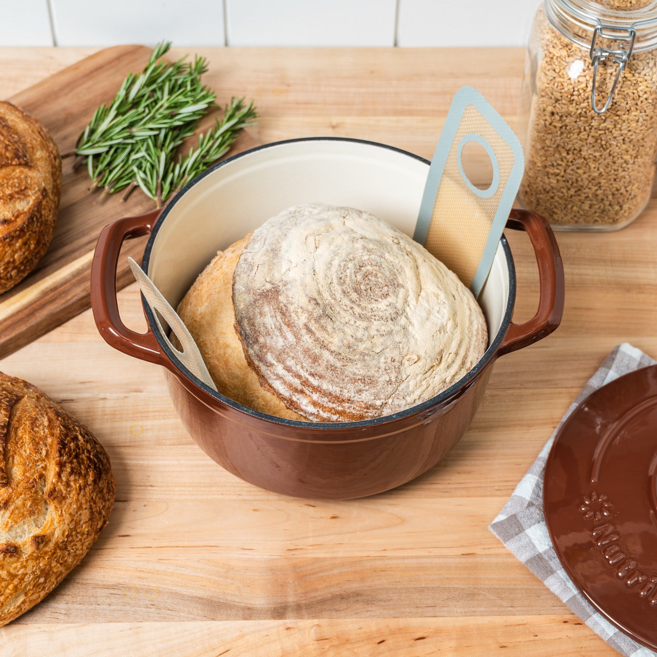 Baked bread in a brown pot on a wooden surface with herbs and grains.