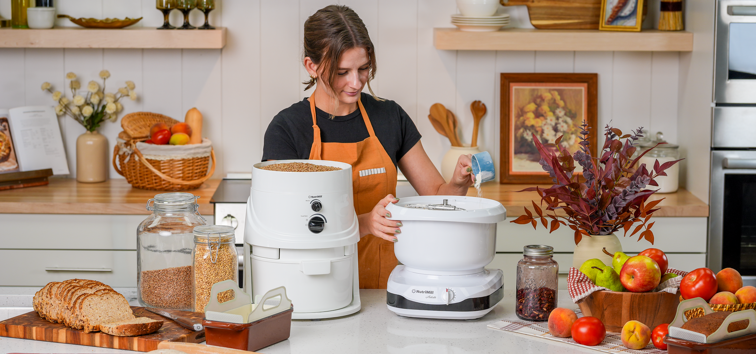 Wide banner view of a woman in an orange apron pouring liquid into the bowl of a white NutriMill Artiste stand mixer beside a white NutriMill grain mill filled with wheat berries, with sliced whole grain bread, glass jars of grain, and a wooden bowl of fresh fruit on the kitchen counter