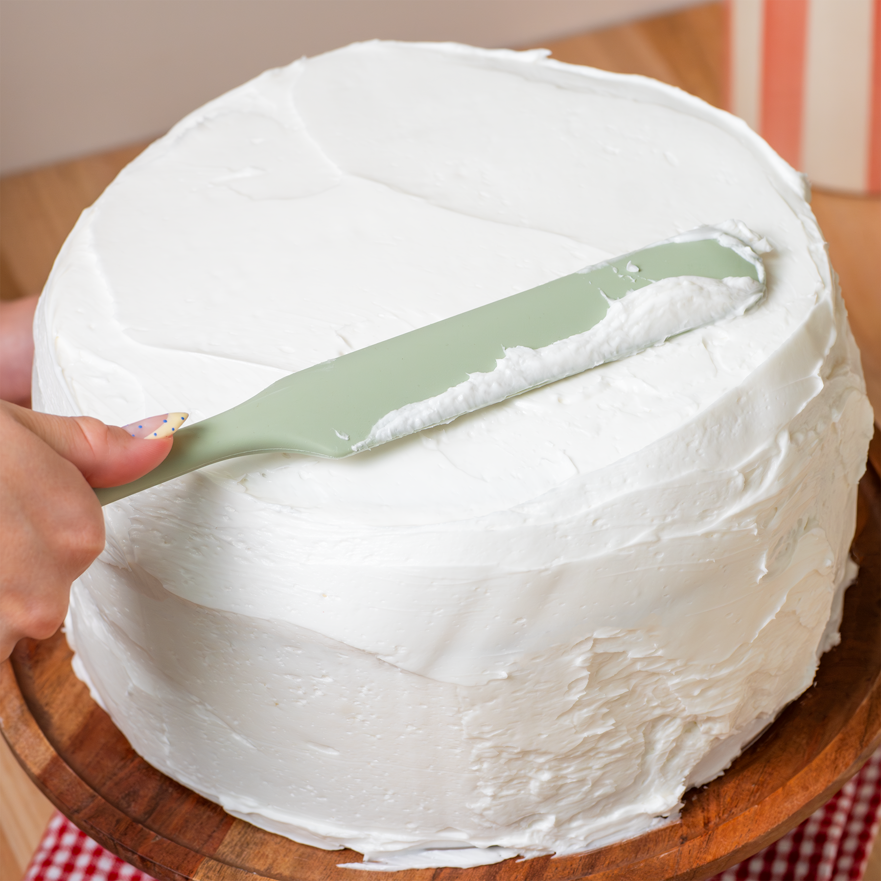 Person frosting a cake with a green silicone spatula on a wooden board.