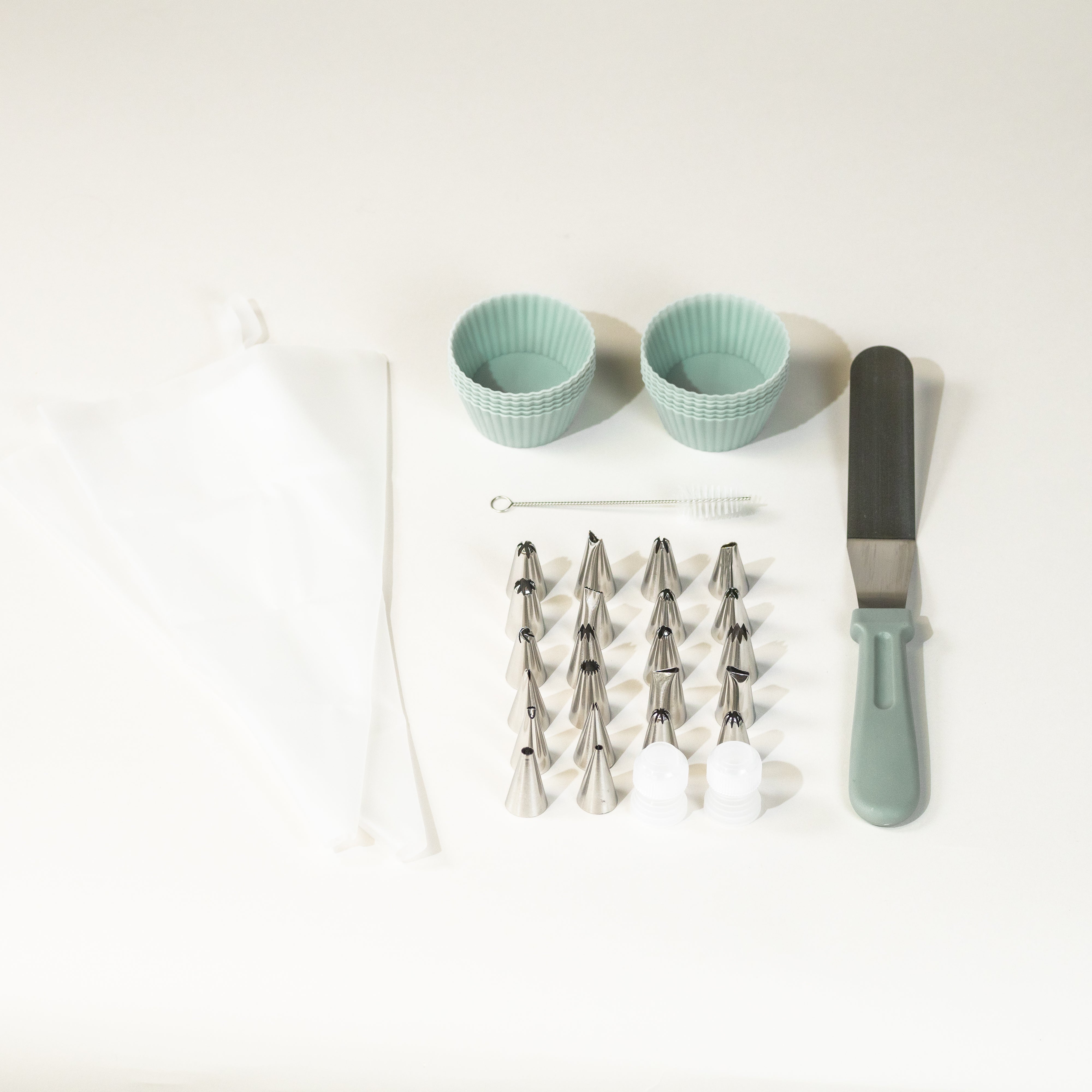A collection of icing tools laid out on a white surface, including set of 12 blue muffin liners, a variety of piping tips, 2 piping bags, decorating tools, and a metal spatula.