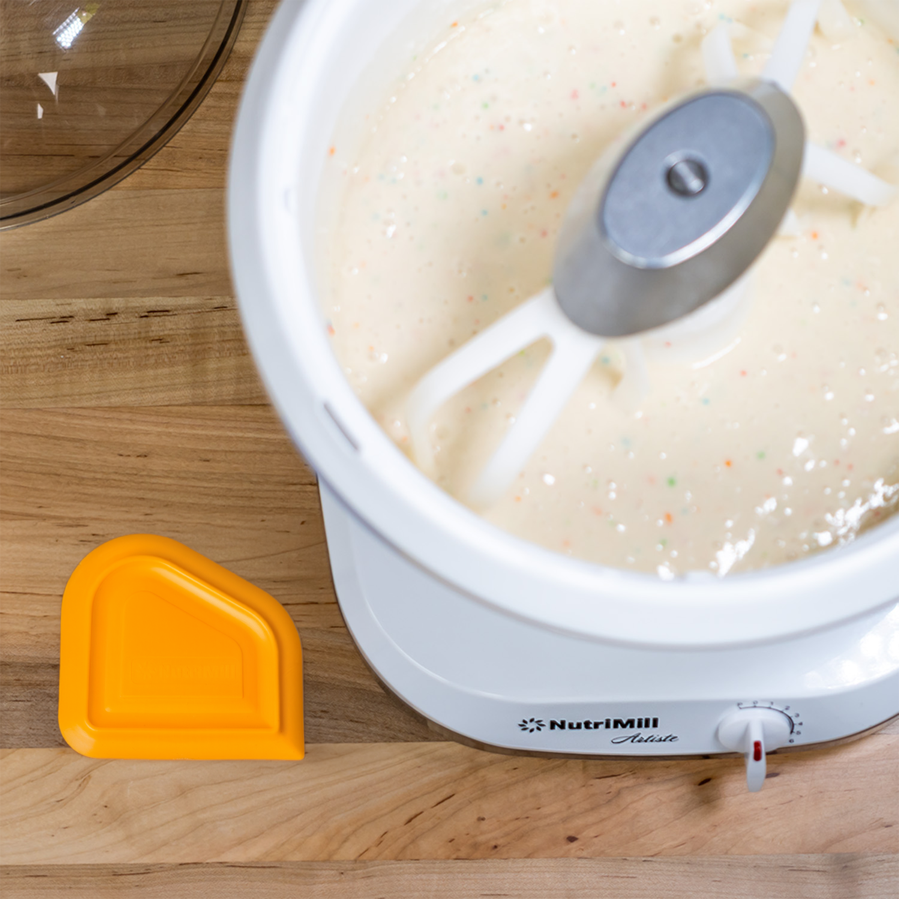 Top view of a NutriMill Artiste mixer bowl filled with funfetti batter next to an orange easy scrape bowl scraper on a wooden counter