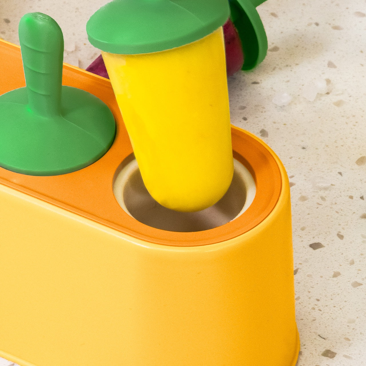Yellow popsicle mold with green popsicle stick and orange a popsicle on a light surface