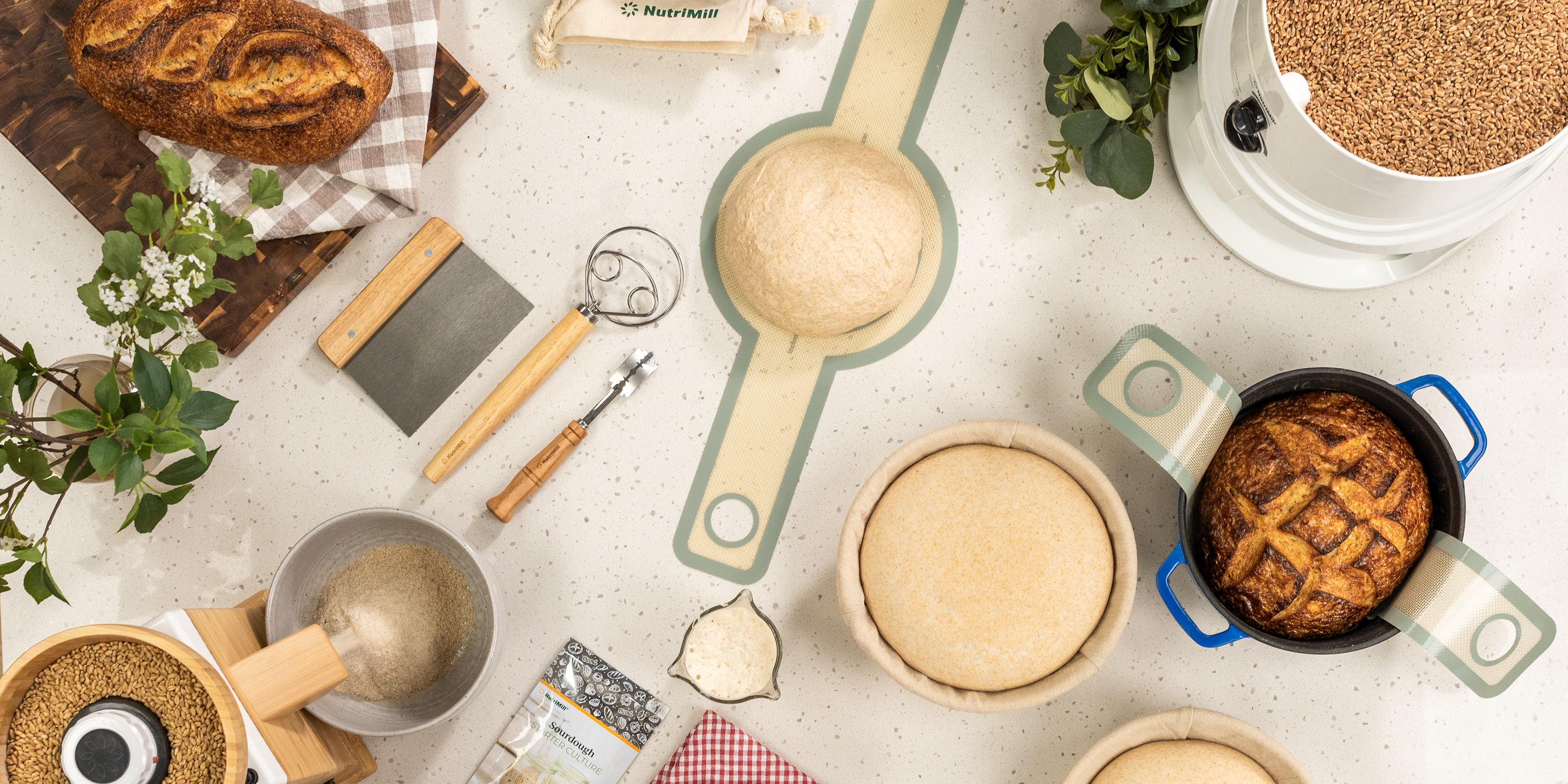 NutriMill grain mills next to bread and the NutriMill sourdough bundle on a white speckled counter