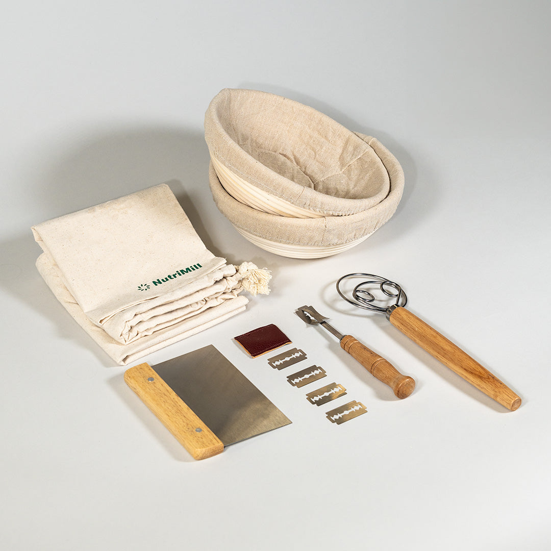 A collection of sourdough tools laid out on a white surface, including 2 banneton baskets with a linen cover, a wooden dough scraper, a metal whisk, a lame with 5 blades, and 2 bread bags.
