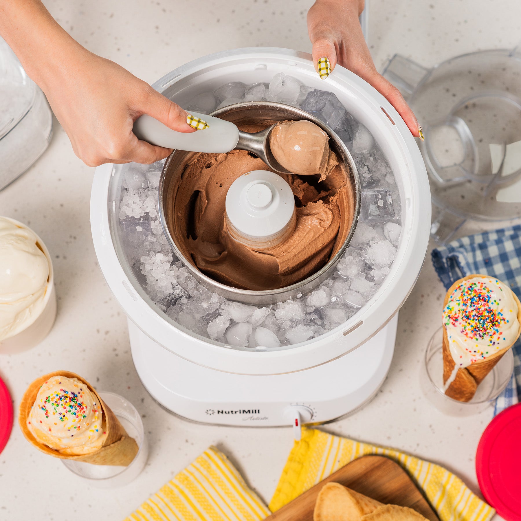 Lifestyle photo of hands using a white ice cream maker with ice cream cones and dishes nearby
