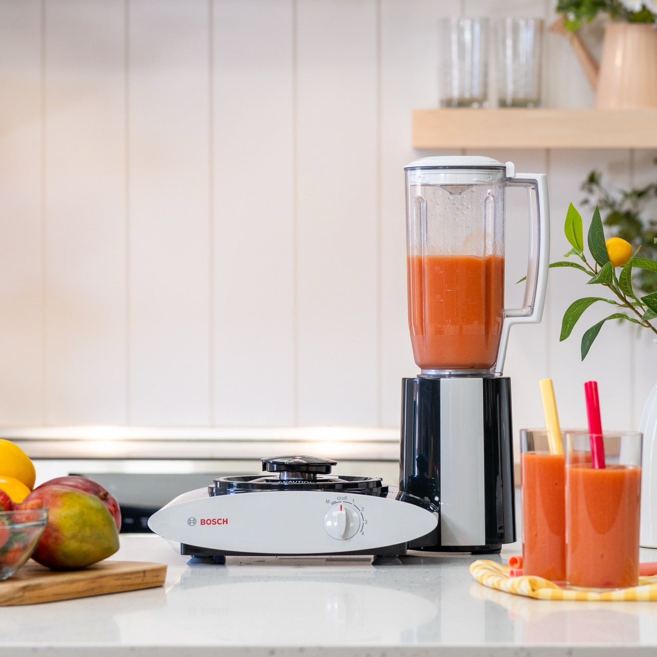 Blender with a glass of juice on a kitchen counter, surrounded by fruits and a Bosch stove.