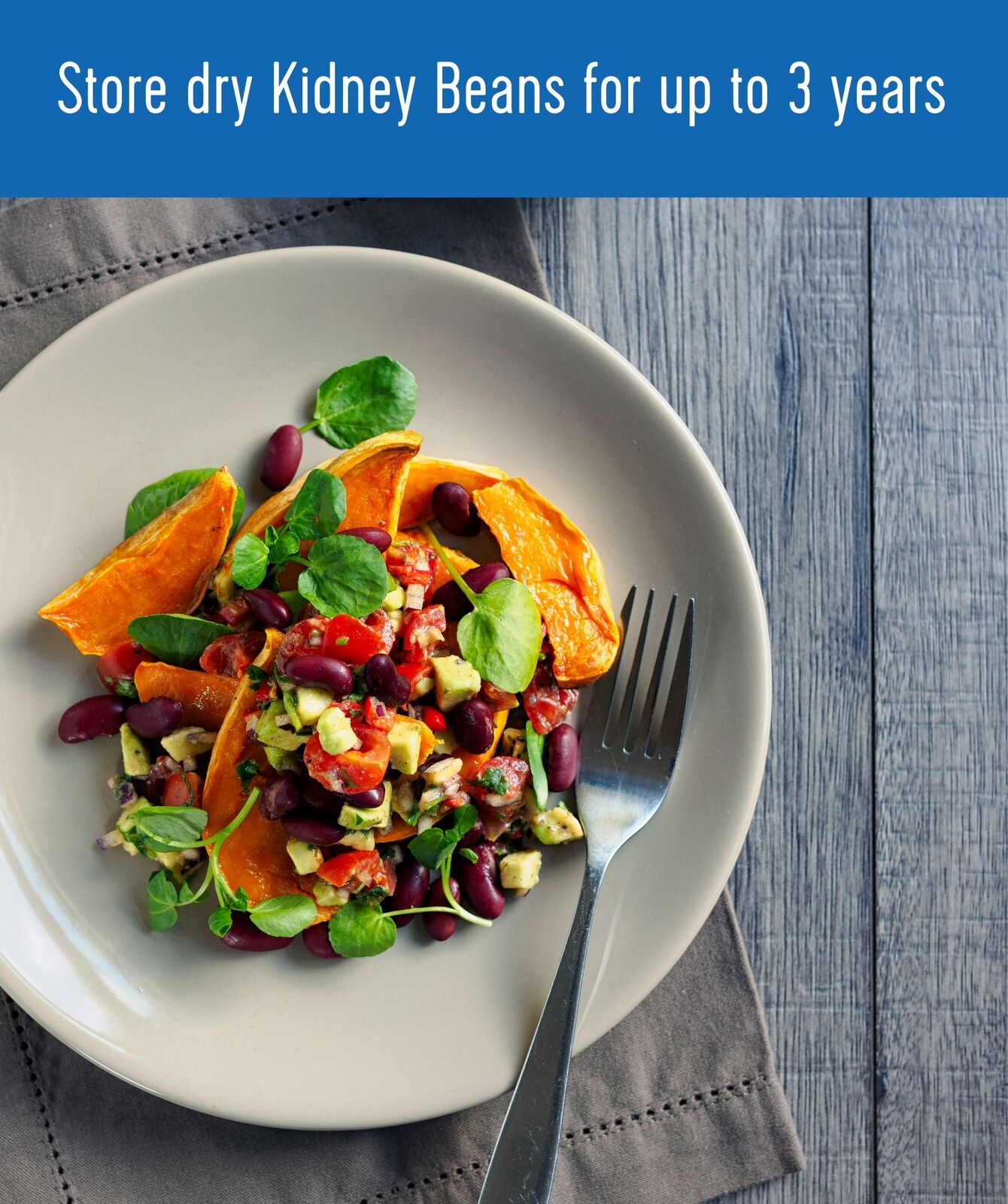Plate of kidney bean salad with sweet potato, avocado, tomato, and watercress with a fork on a grey napkin and wood surface, with text 'Store dry Kidney Beans for up to 3 years'
