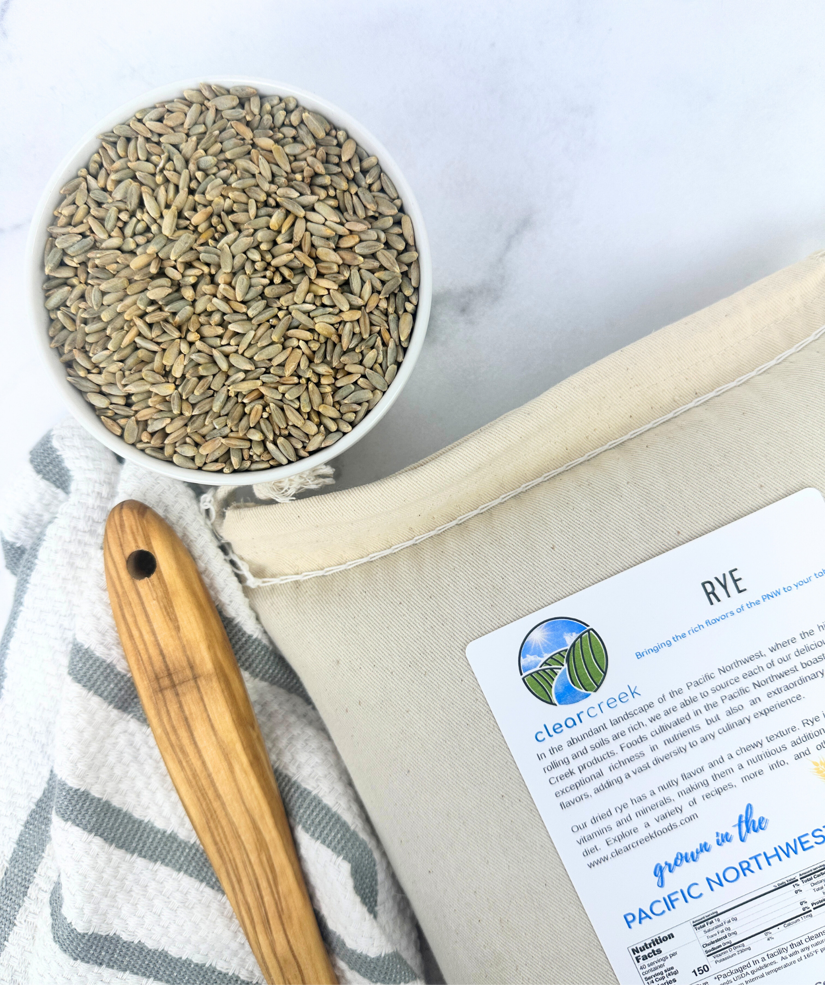 Clear Creek Rye canvas bag beside a bowl of rye berries with a wooden spoon on a marble surface