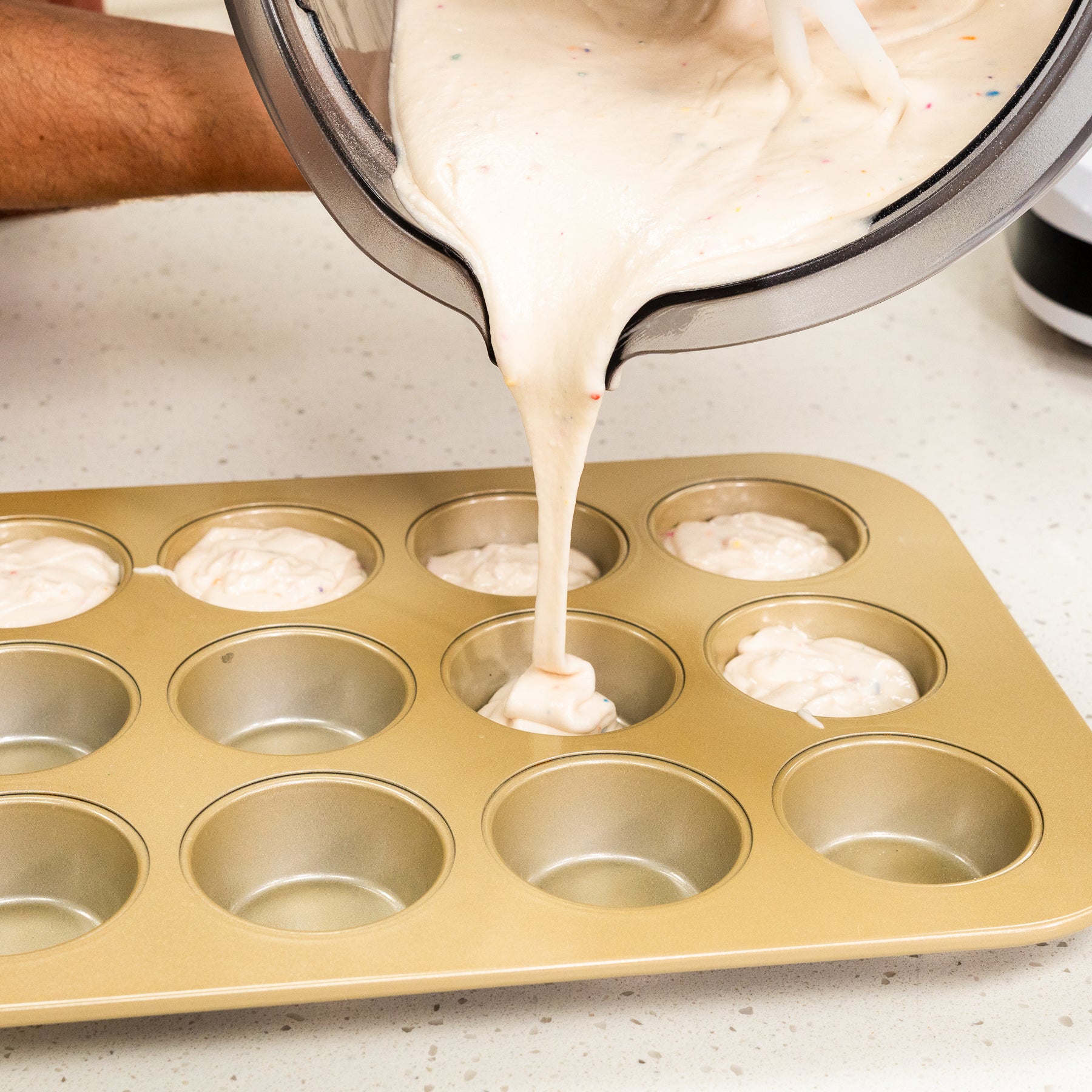 Person pouring white batter with sprinkles into gold muffin tin on white speckled counter