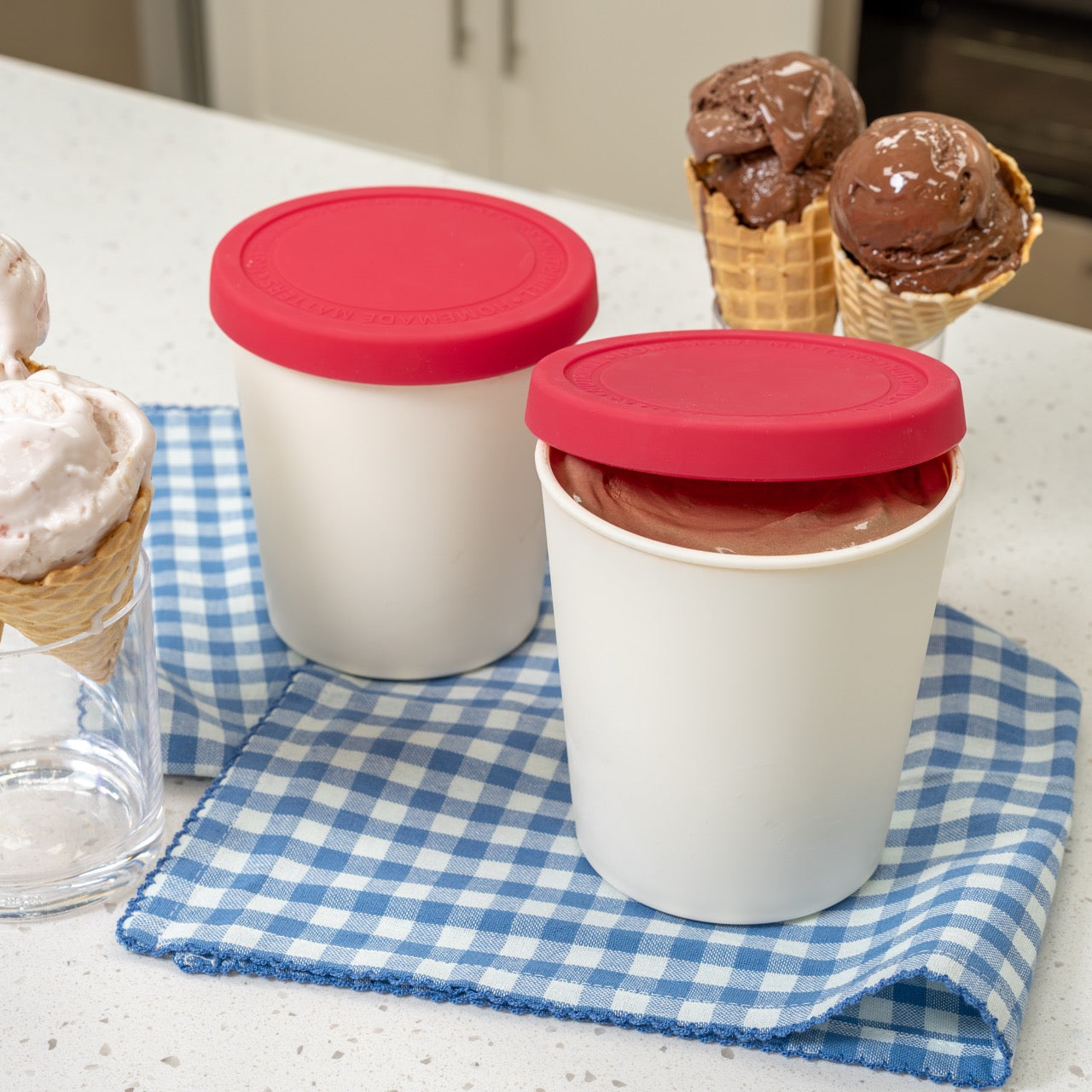 Two containers with red lids filled with ice cream on a checkered cloth, with ice cream cones in the background.