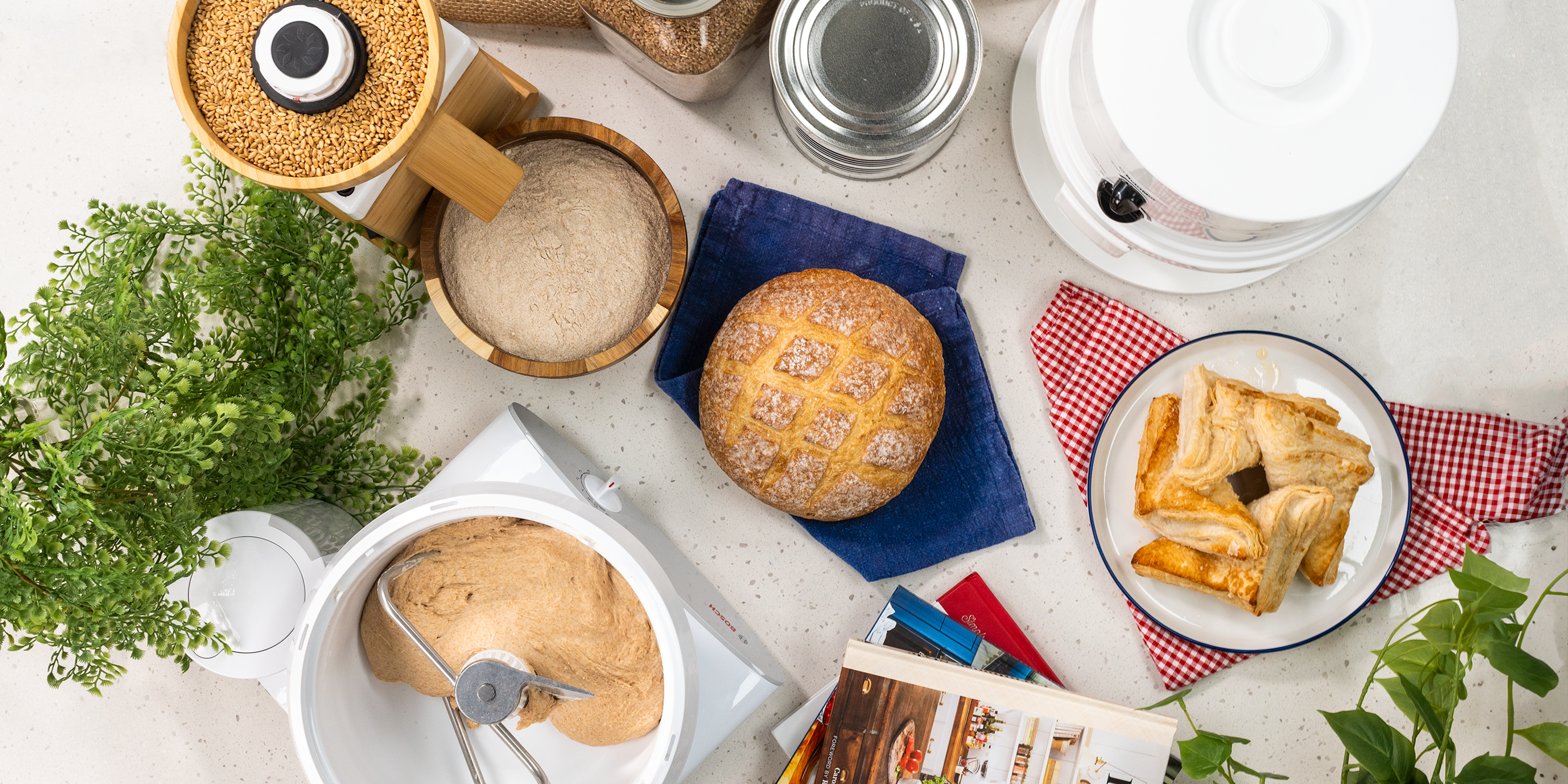 Various Bosch and NutriMill kitchen appliances on a white speckled counter next to sourdough bread, a dessert pastry, and decorations