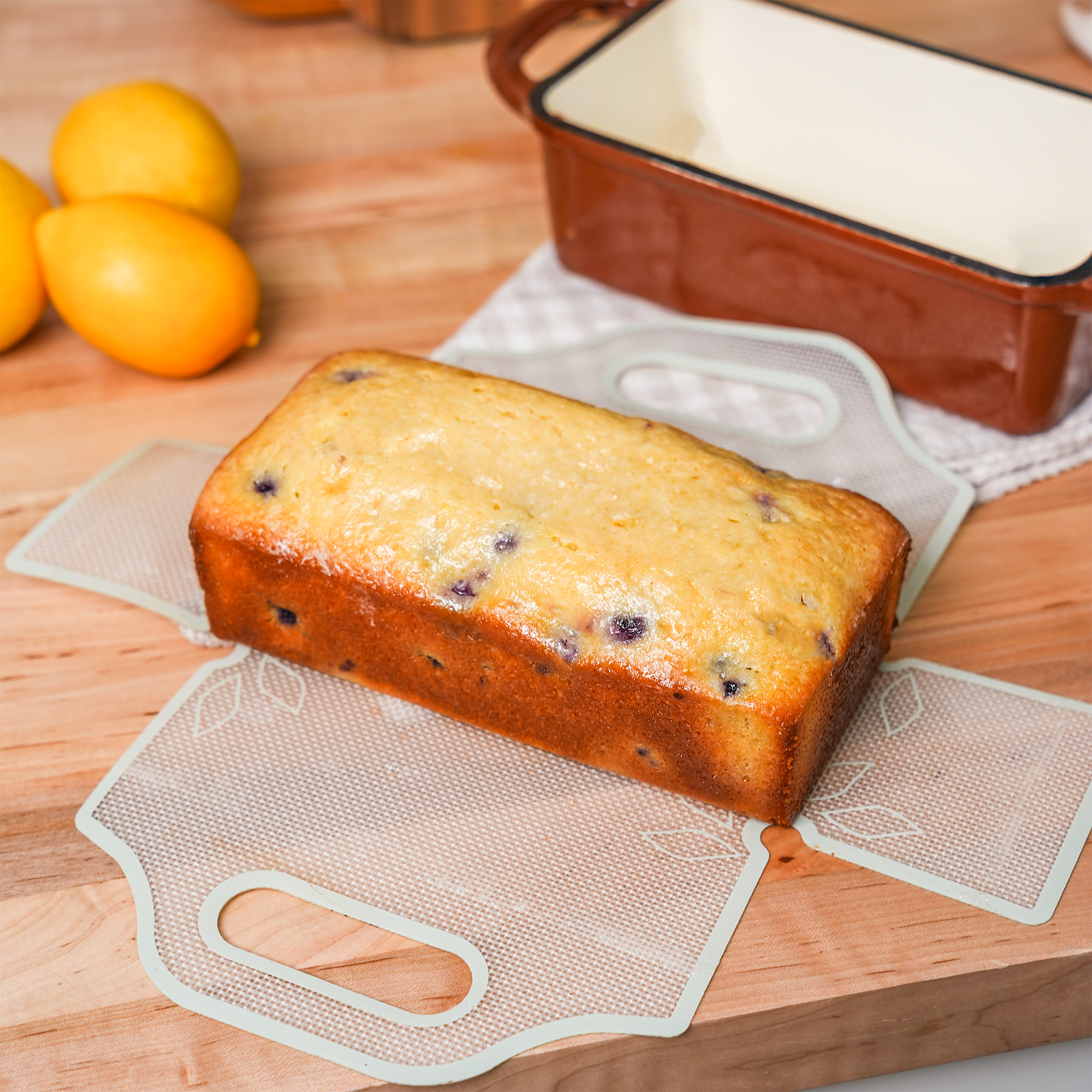 Lemon blueberry loaf on a parchment paper sling beside a ceramic bread pan and lemons