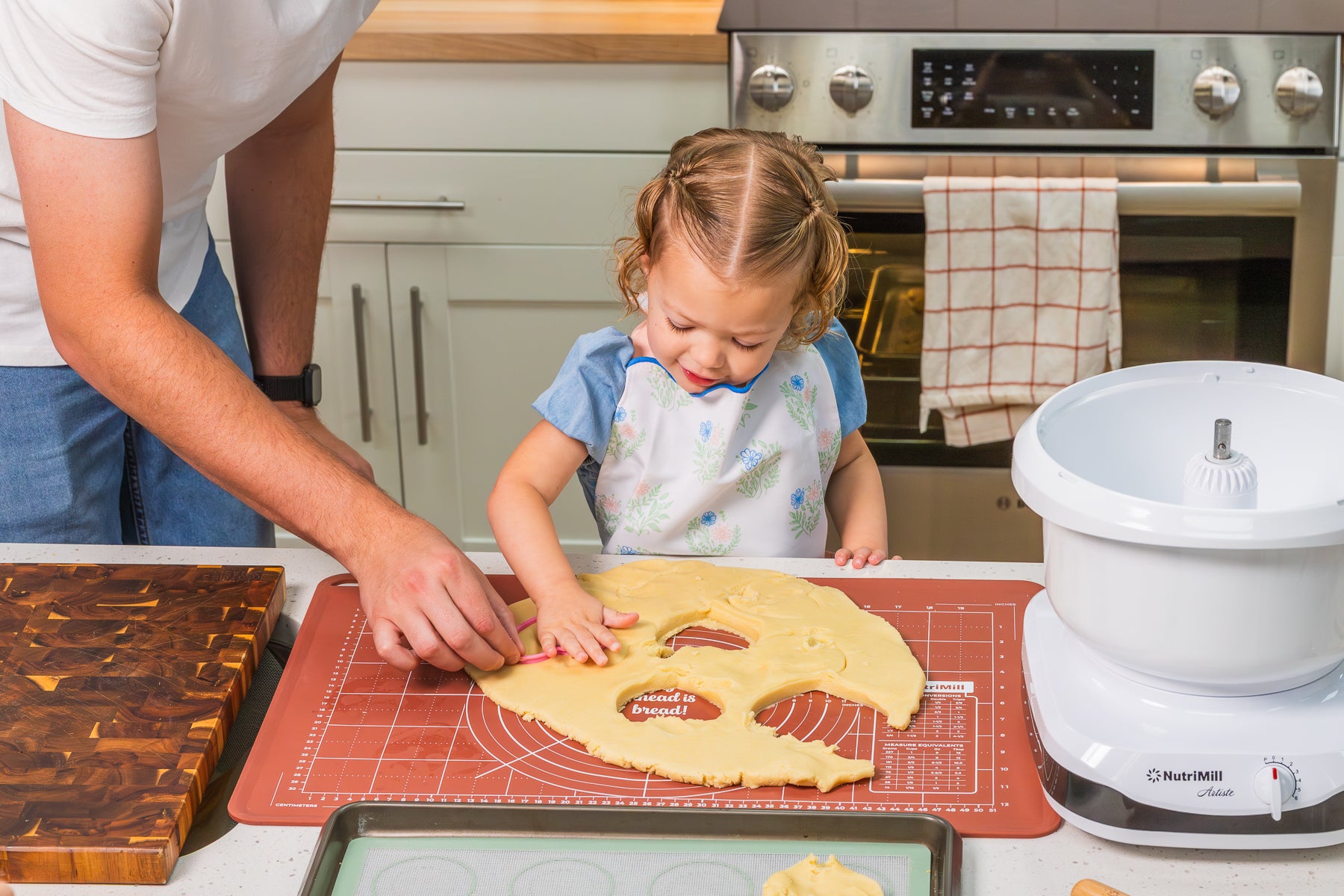 Toddler girl cutting out cookies from rolled dough on a NutriMill pastry mat with help from an adult, beside a white NutriMill Artiste mixer