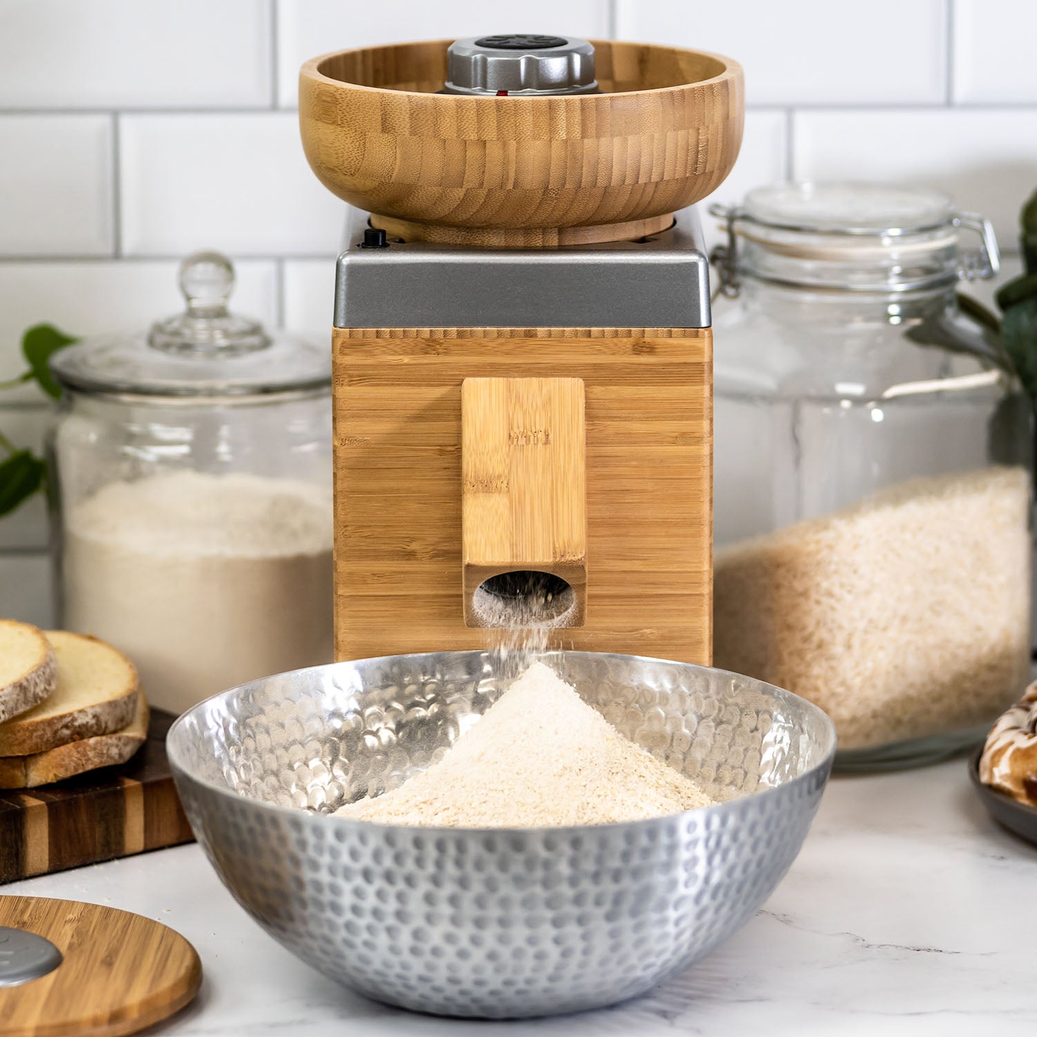 A NutriMill harvest grain mill with a silver trim milling grains into a silver bowl with grains and a white wall in the background