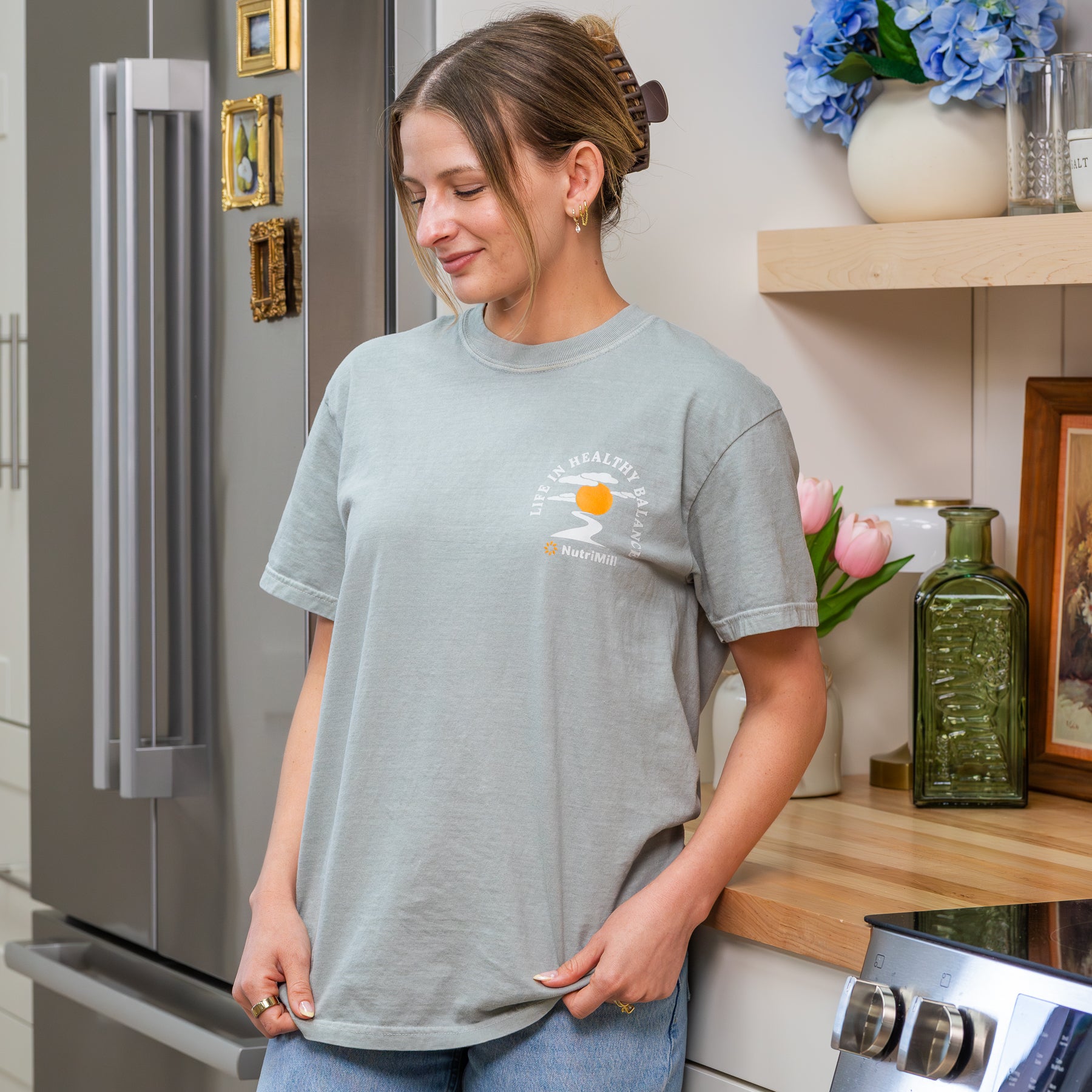 Woman wearing a sage green NutriMill 'Life in Healthy Balance' t-shirt with sunset pocket logo looking down in a kitchen