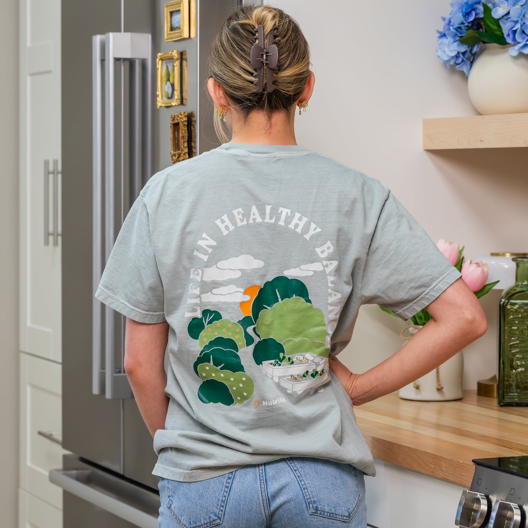 Back view of a woman wearing a sage green NutriMill 'Life in Healthy Balance' t-shirt featuring a garden landscape illustration