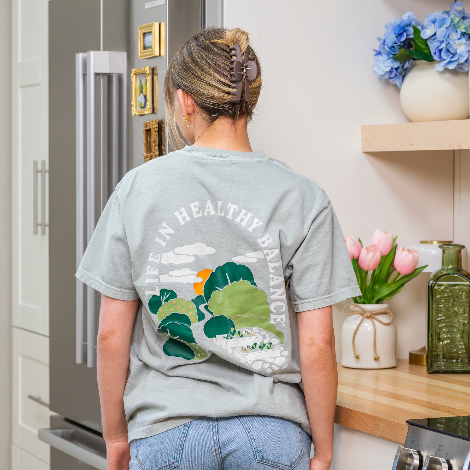 Back view of a woman wearing a sage green NutriMill 'Life in Healthy Balance' t-shirt with garden landscape illustration in a kitchen