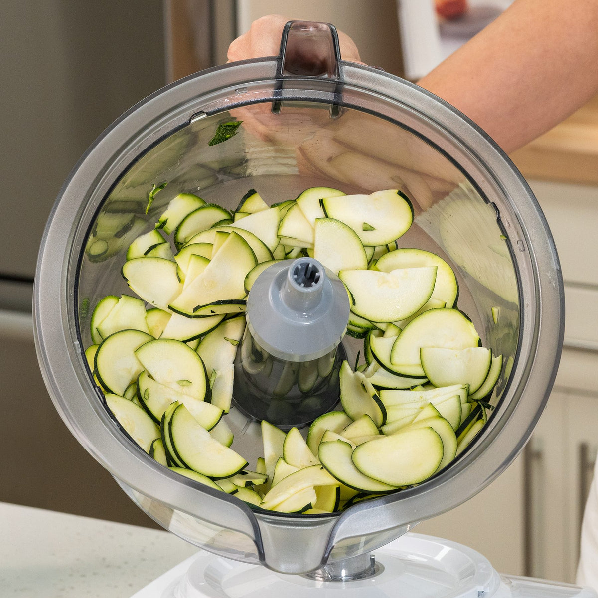 Hand tilting a clear mixing bowl with a spout and handle, filled with thinly sliced zucchini