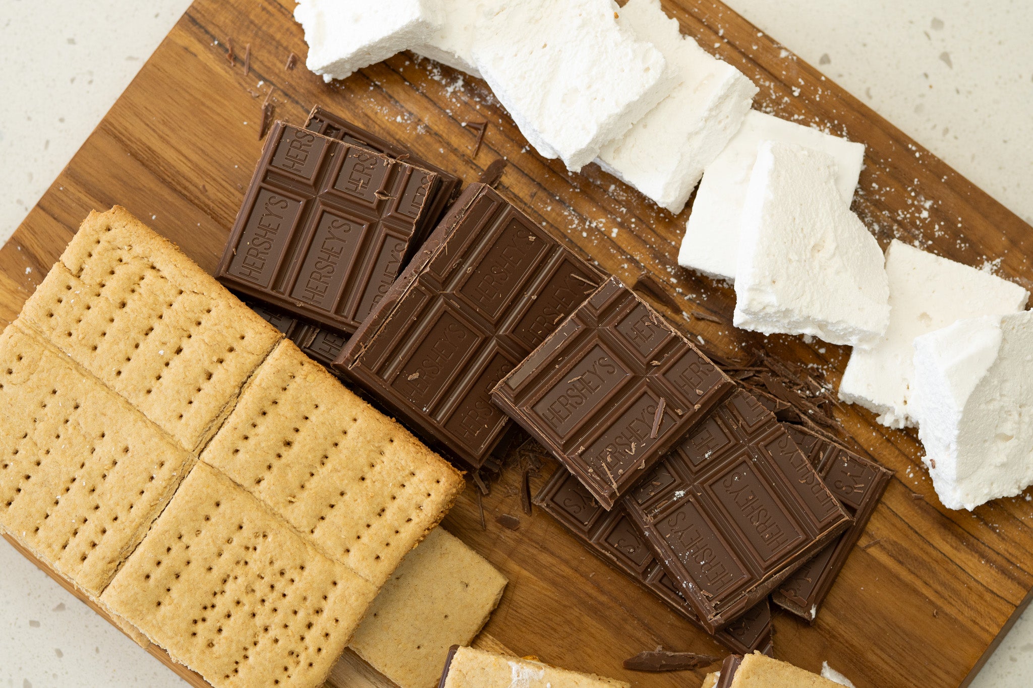 S'mores ingredients arranged on a wooden cutting board with stacked Hershey's chocolate bars, graham crackers, and homemade marshmallows