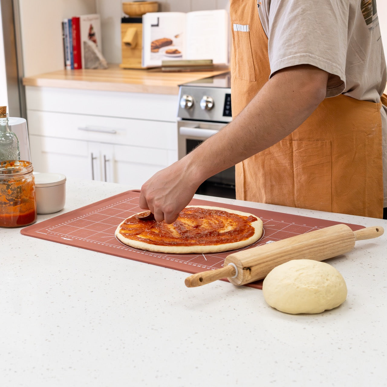 Person making a pizza in a kitchen with a rolling pin and dough mat on a counter.