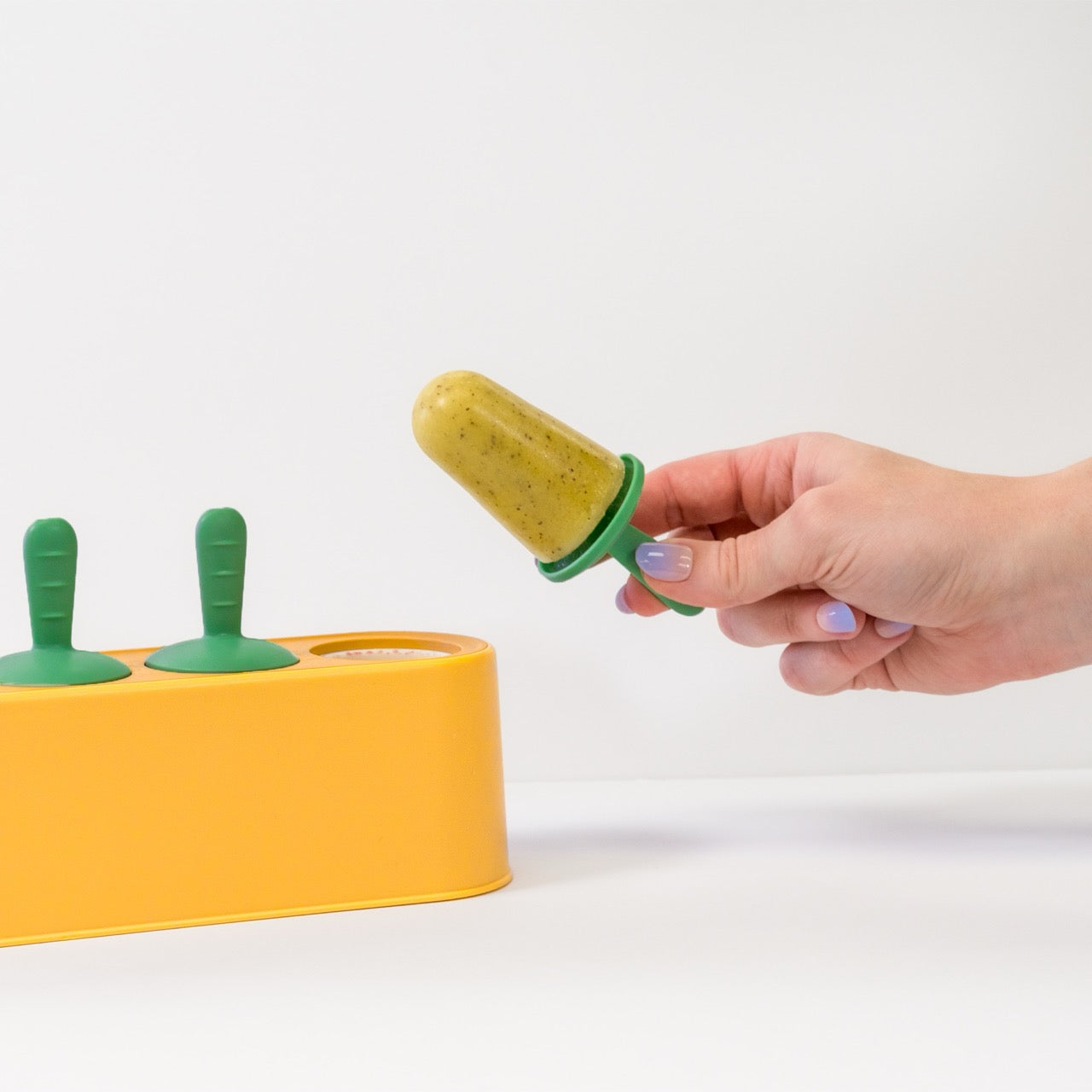 Hand holding a popsicle with a yellow base and green handles, next to a yellow popsicle mold.