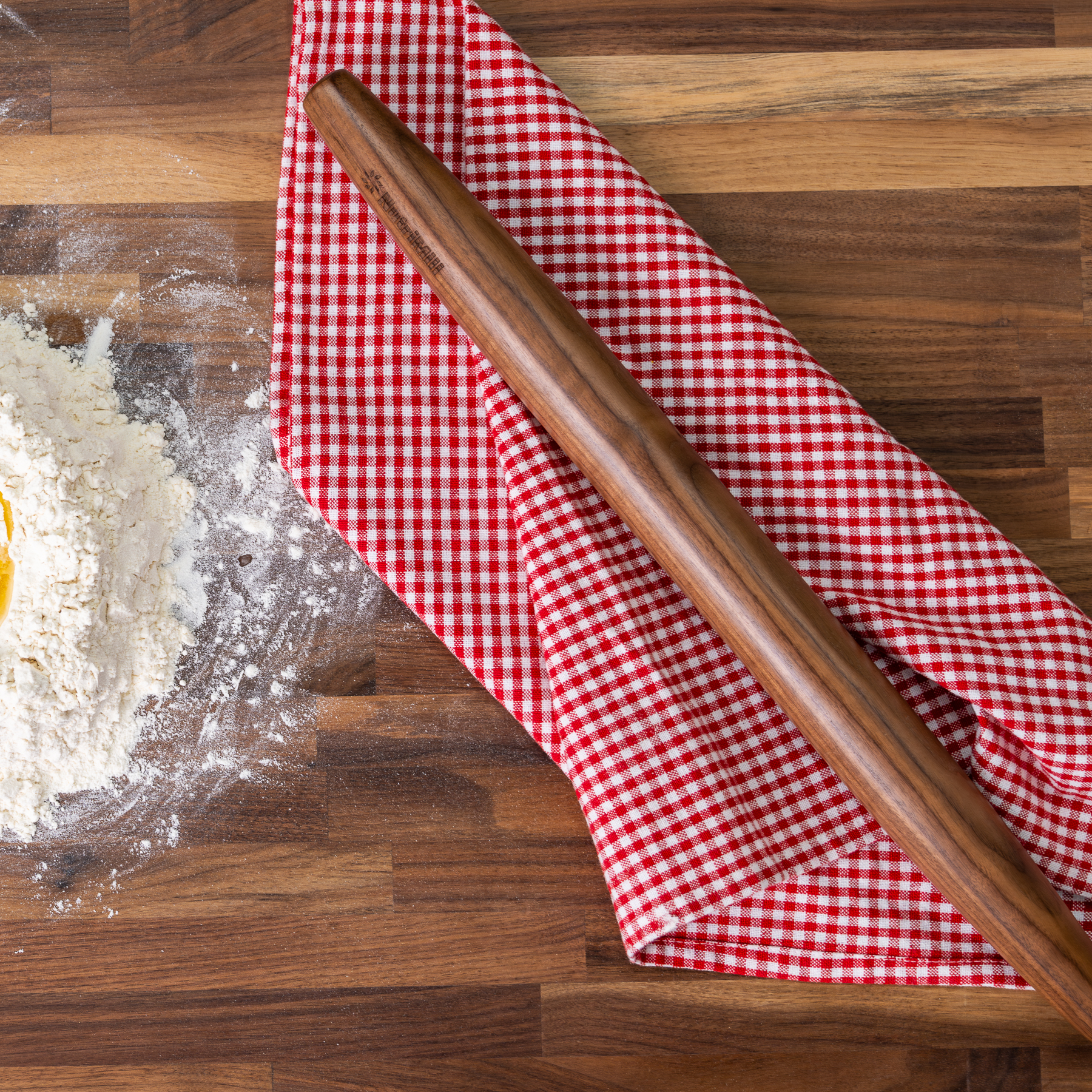 Top view of the NutriMill walnut French rolling pin on a red gingham kitchen towel next to a mound of flour on a butcher block counter
