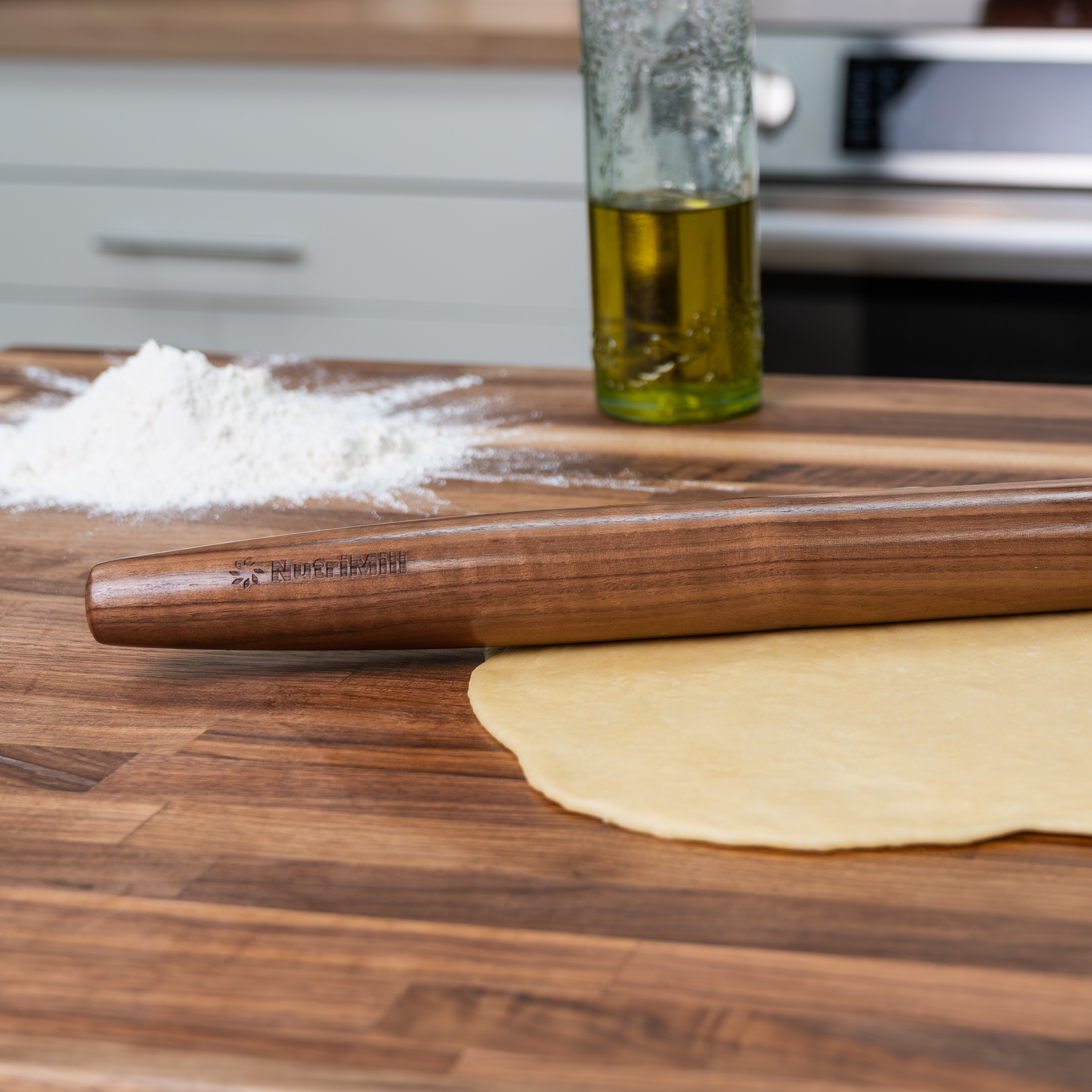 NutriMill walnut French rolling pin on a butcher block counter beside rolled-out dough, with a mound of flour and a bottle of olive oil in a bright kitchen