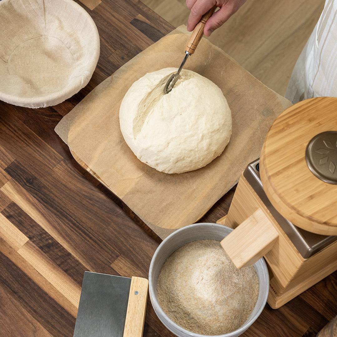 Hand scoring a sourdough boule on parchment paper next to a NutriMill grain mill, banneton basket, and dough scraper on a wooden counter