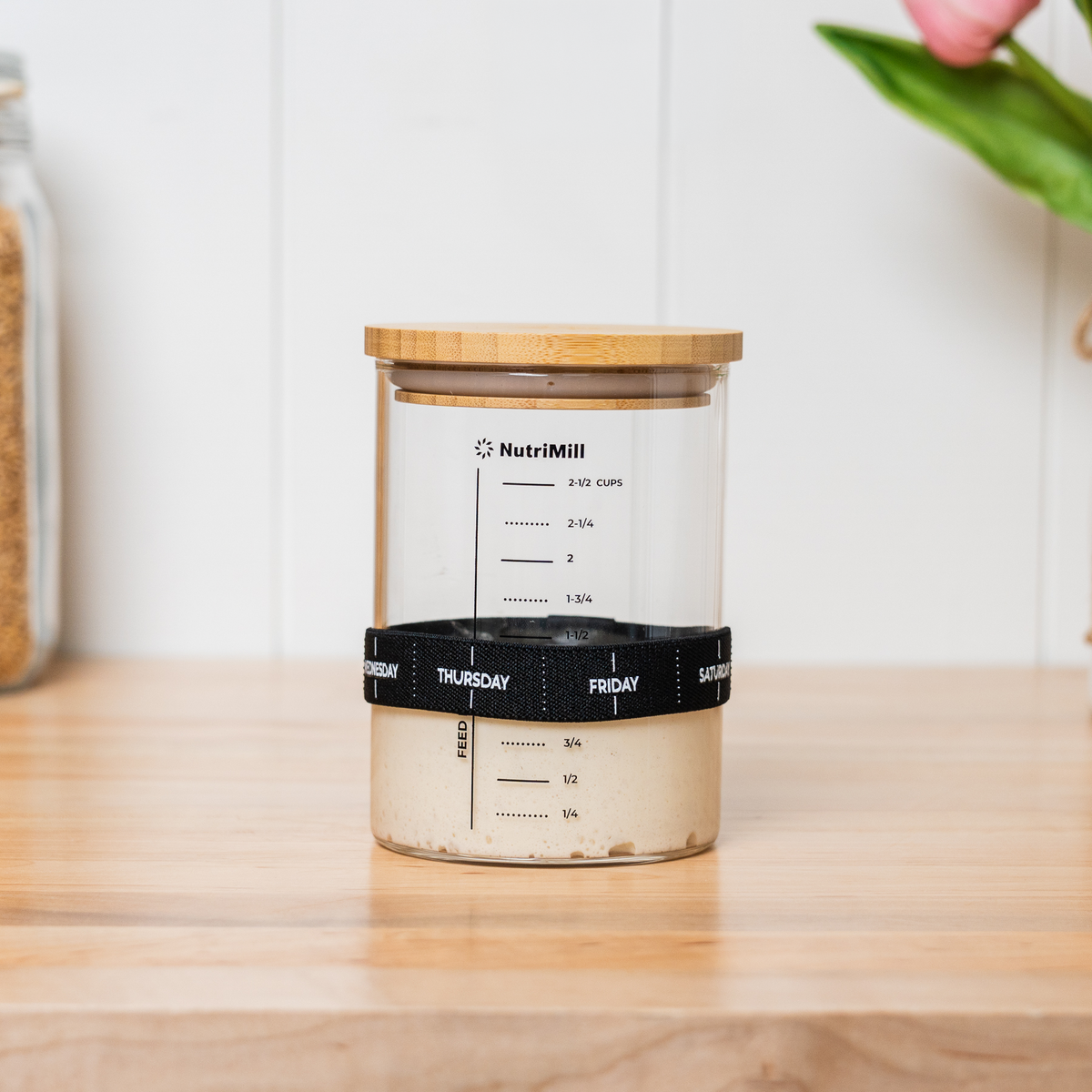 NutriMill 2½-cup sourdough starter jar with bamboo lid and feeding band set to Thursday, filled with starter, on a wood countertop with tulips in the background