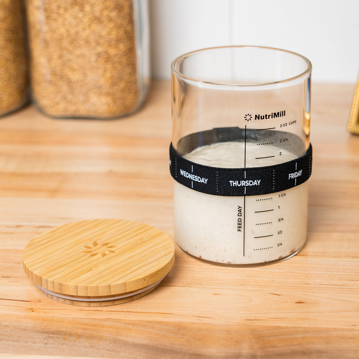 NutriMill sourdough starter jar open with its bamboo lid resting on the wood countertop in front of it, starter inside and feeding band set to Thursday, jars of grain in the background