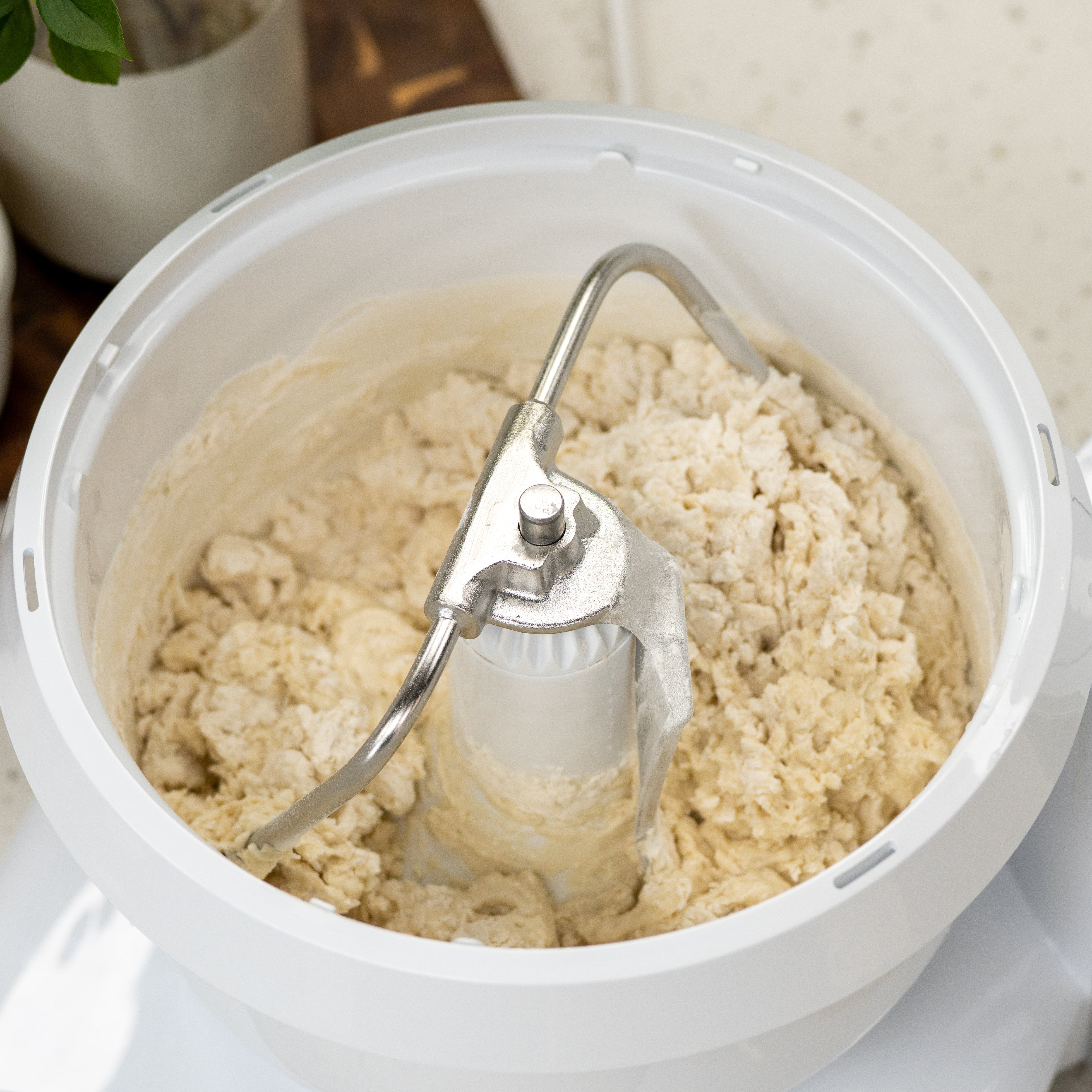 Stainless steel dough hook kneading bread dough inside a white Bosch Universal Plus mixer bowl