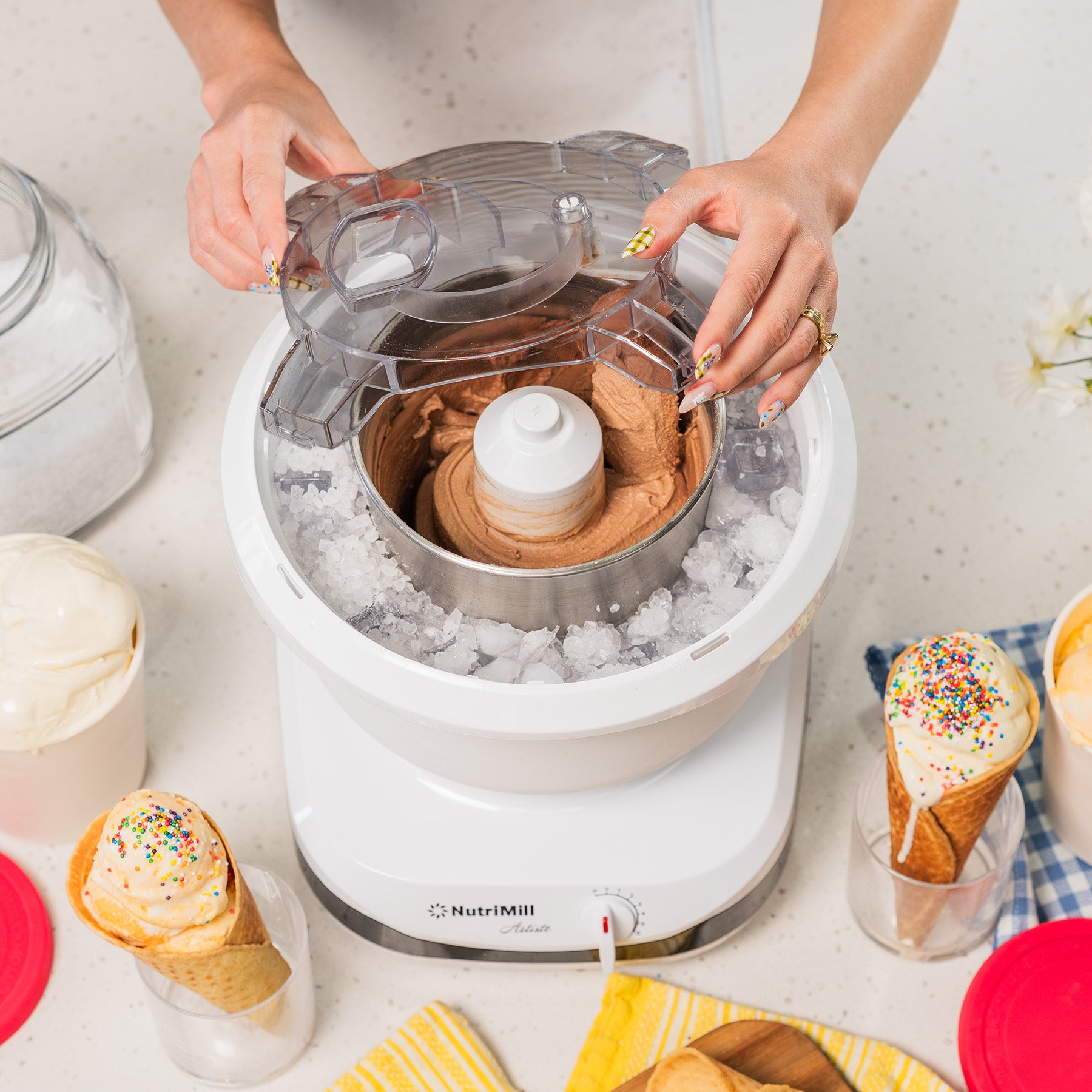 Lifestyle photo of hands using a white ice cream maker with ice cream cones and dishes nearby