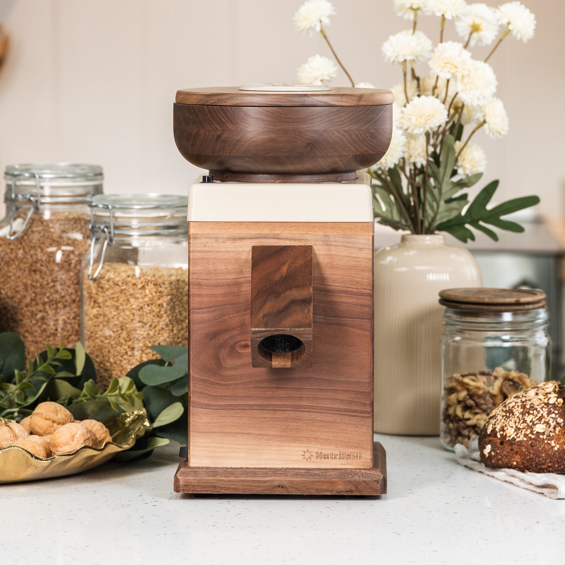 NutriMill Walnut harvest grain mill with cream-colored trim on white counter with plants and grains in background
