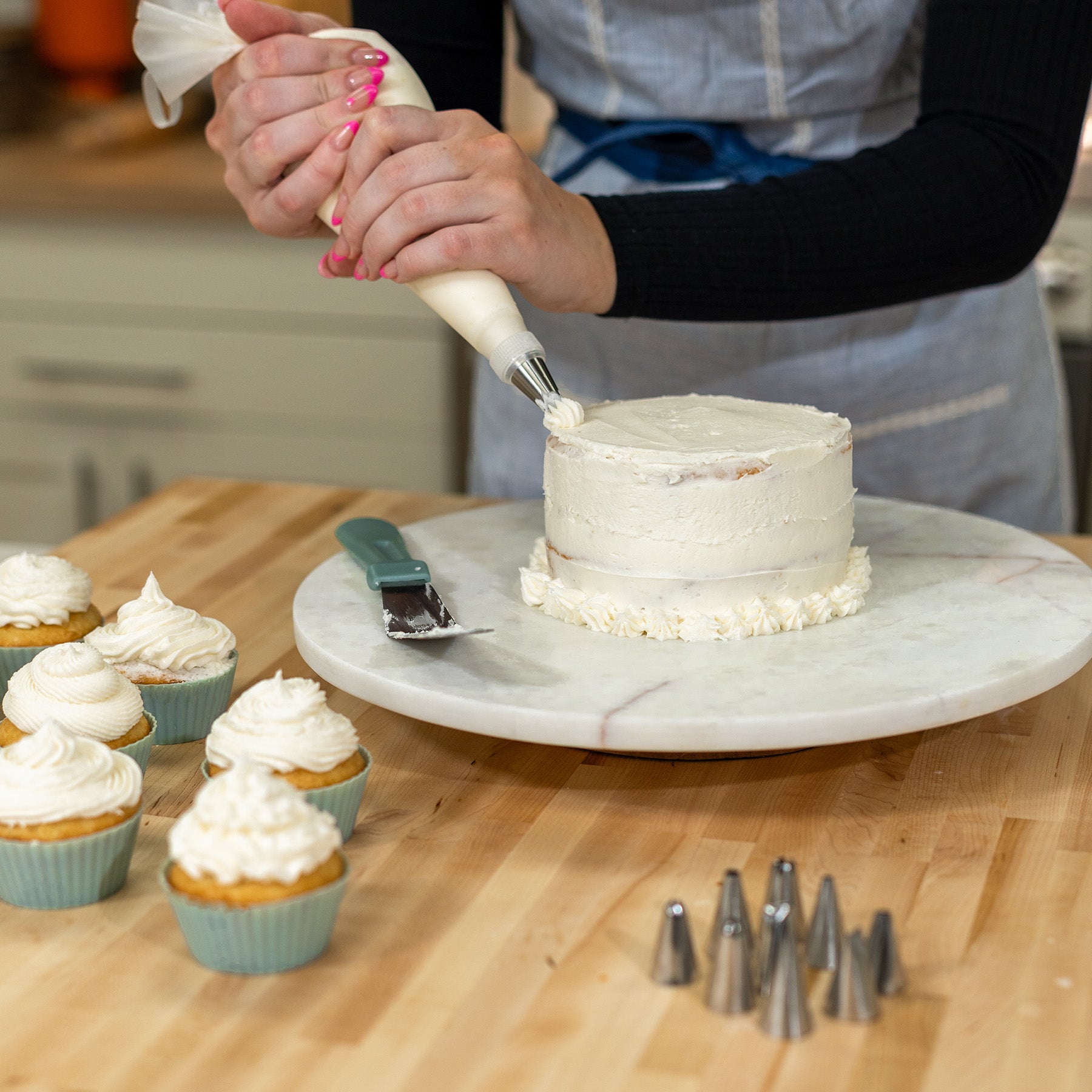 Woman icing a white cake on a white platter on a wooden counter next to vanilla cupcakes