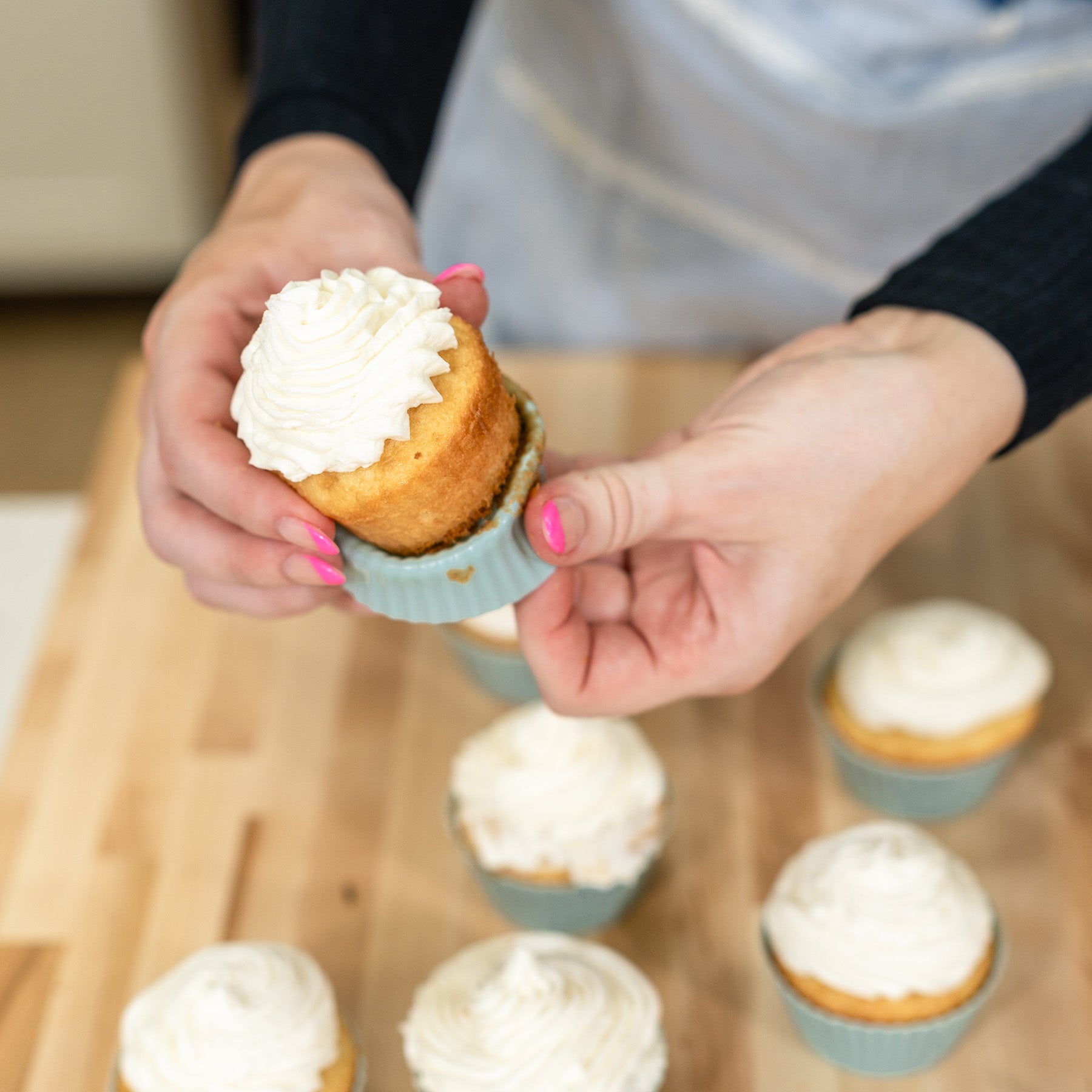 A woman peeling back the cupcake liner on a vanilla cupcake with more cupcakes in the background
