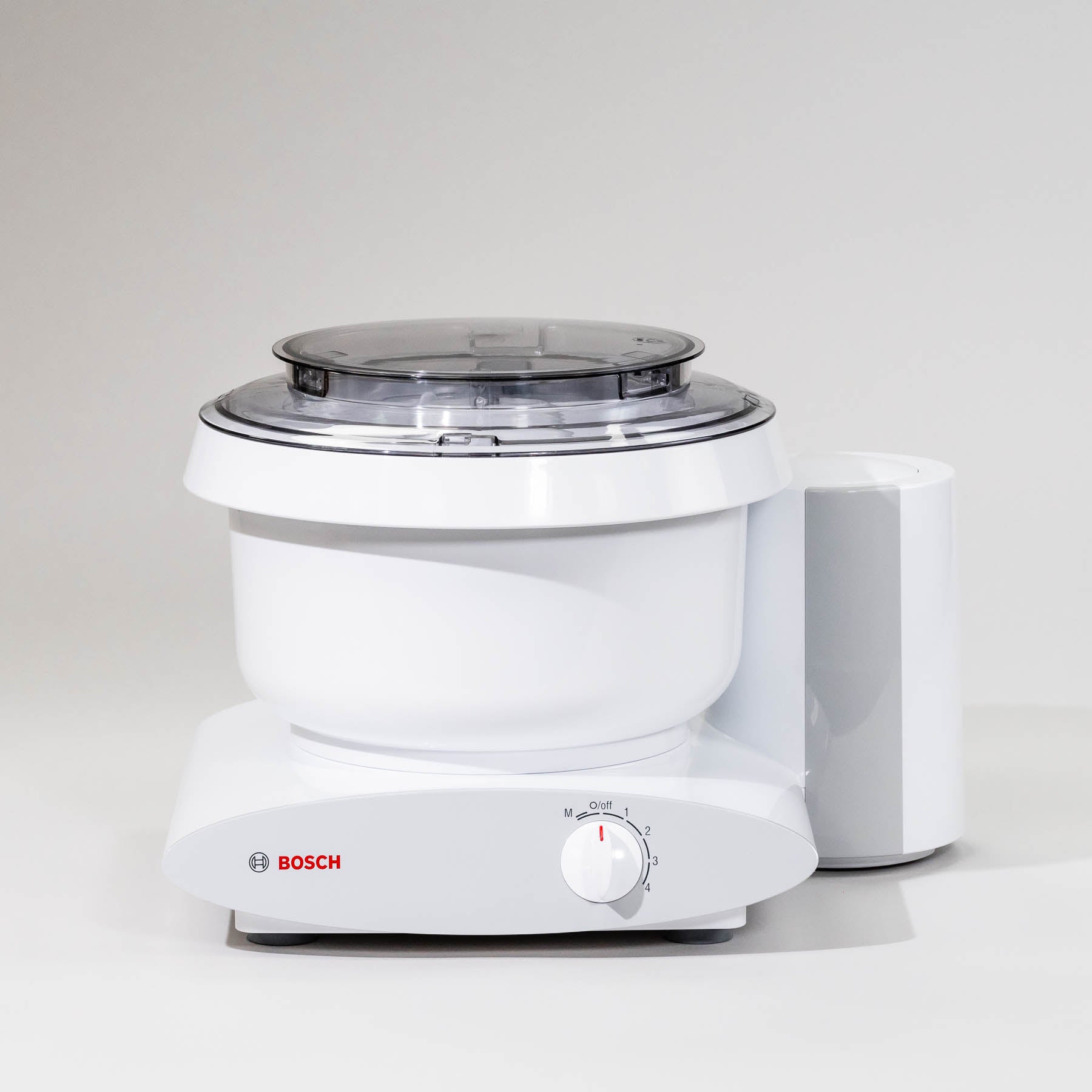 A white Bosch Universal Plus mixer with a clear lid, featuring a speed control knob, suction feet on the base, and a dough hook attachment.