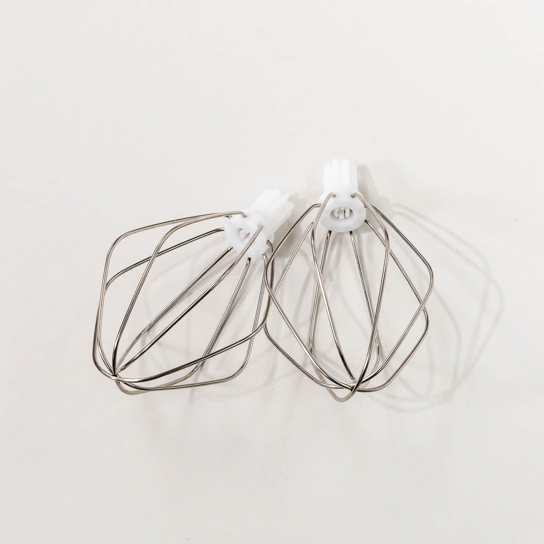 Two stainless steel wire whisks for a Bosch mixer placed on a white surface.
