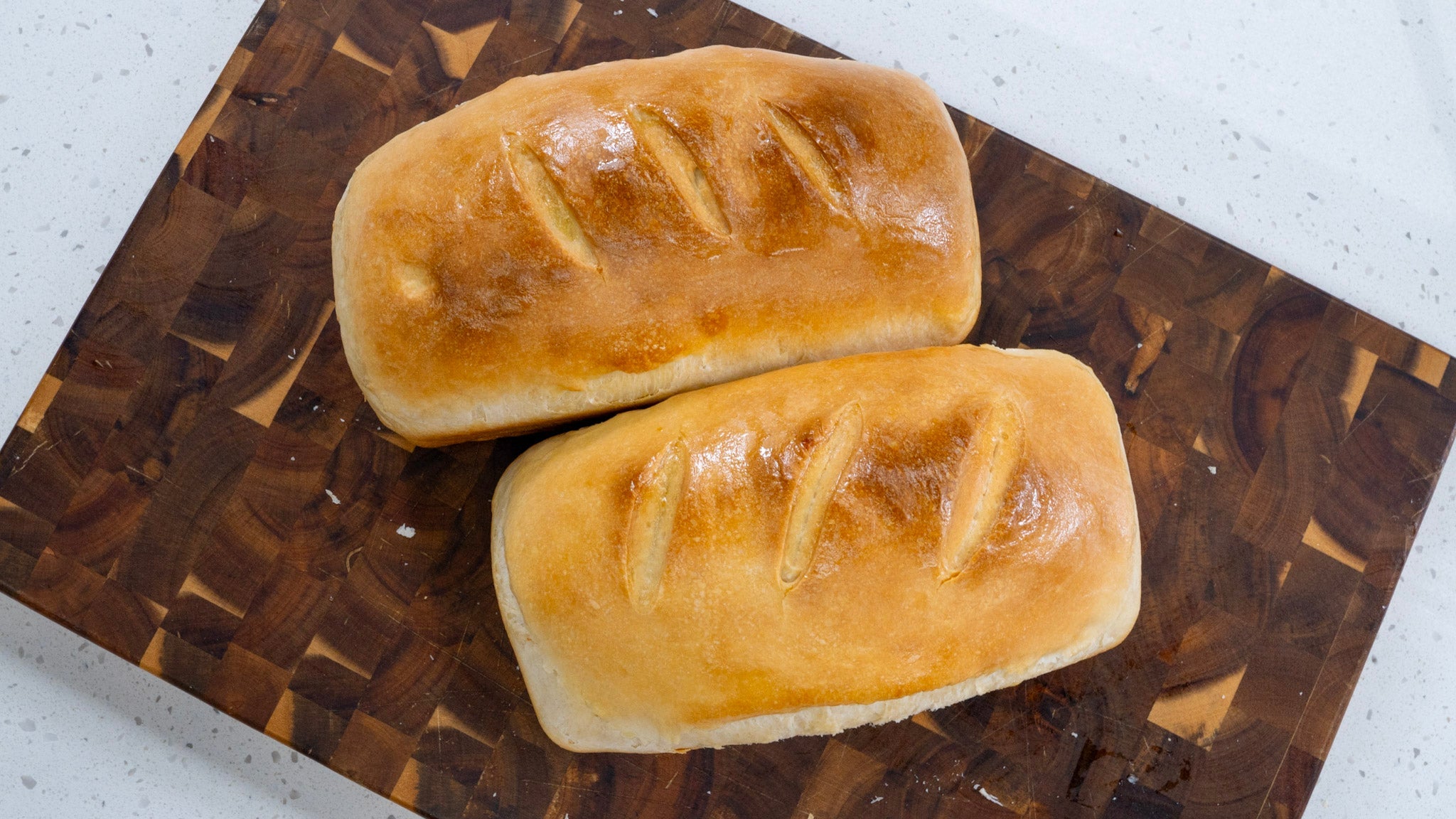 Two golden-brown loaves of sourdough sandwich bread with three decorative slashes each, cooling on a dark wooden cutting board.