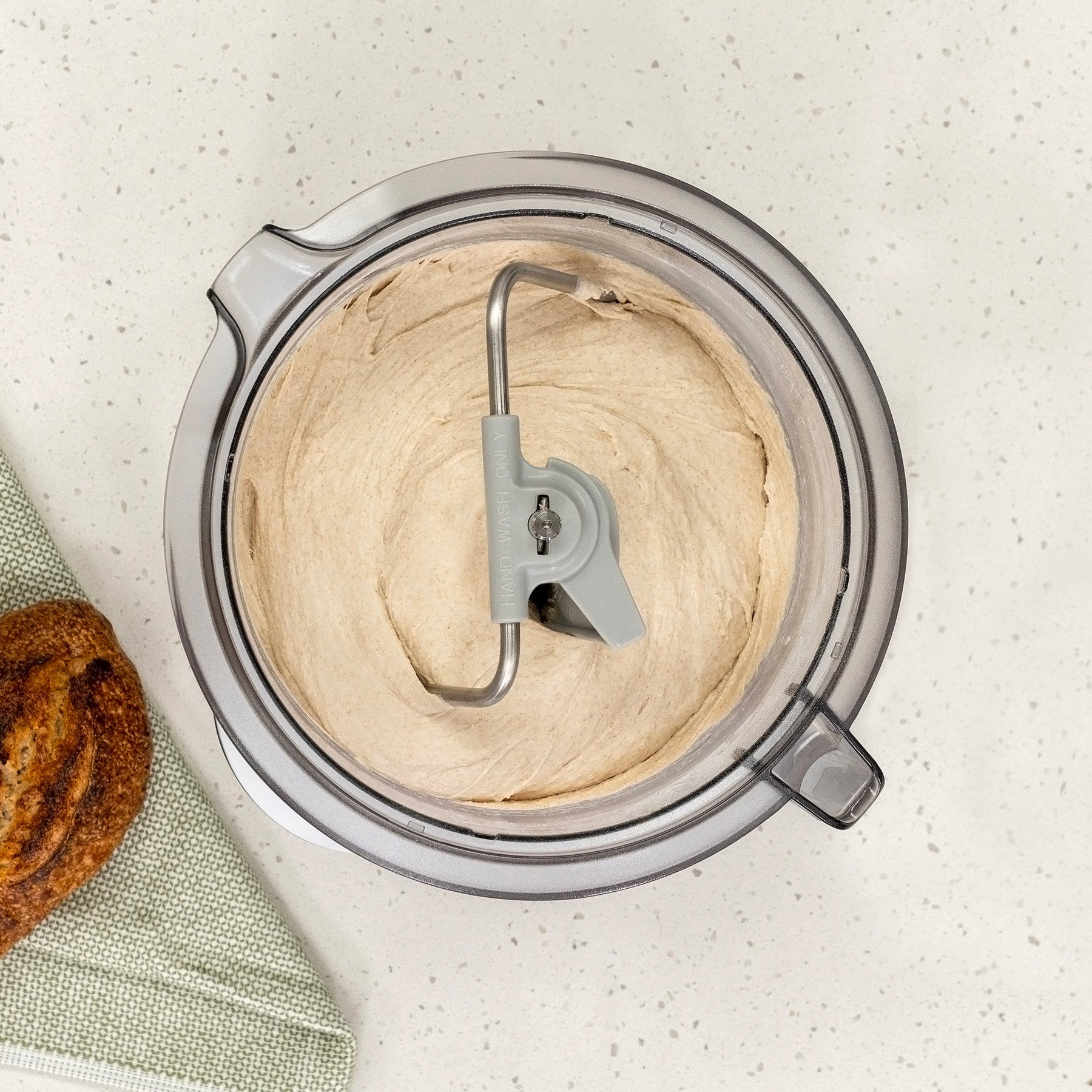 Bird's eye view of white batter with sprinkles in clear bowl with metal dough hook attached, sitting on a white speckled counter