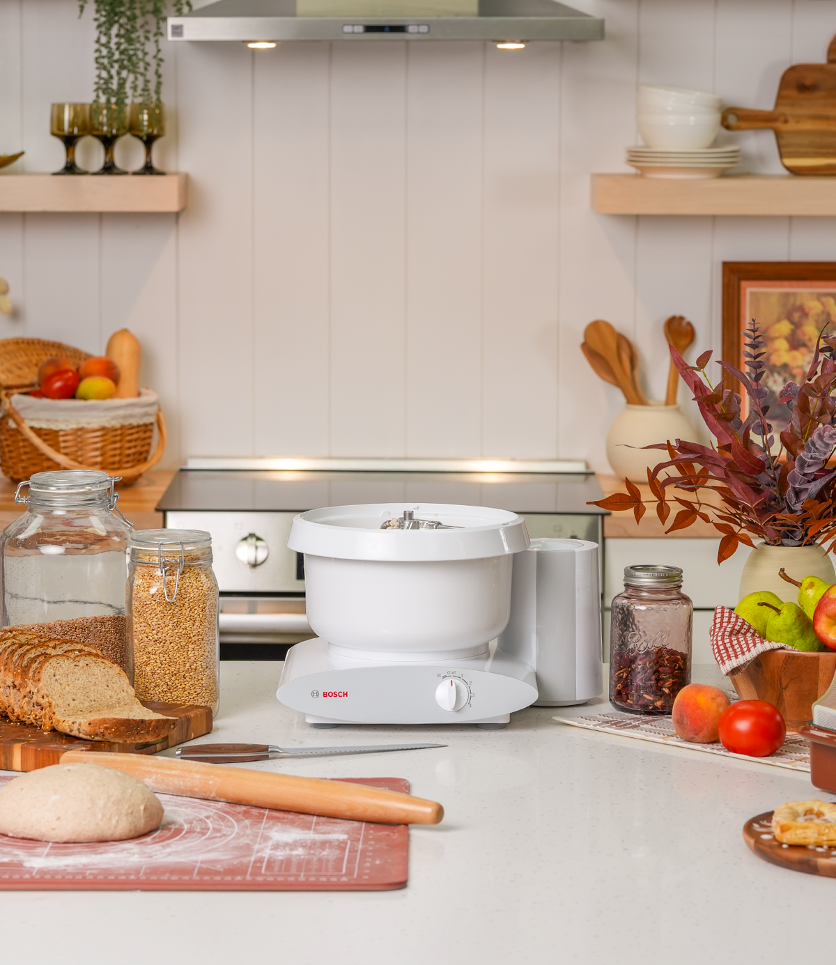 Autumn kitchen scene in a taller crop centered on a white Bosch Universal Plus stand mixer, with a stainless range hood and open wood shelving above, a basket of bread and glass jars of flour and wheat berries at left, and a jar of dried berries, a bowl of apples and pears, and a vase of dried fall foliage at right.