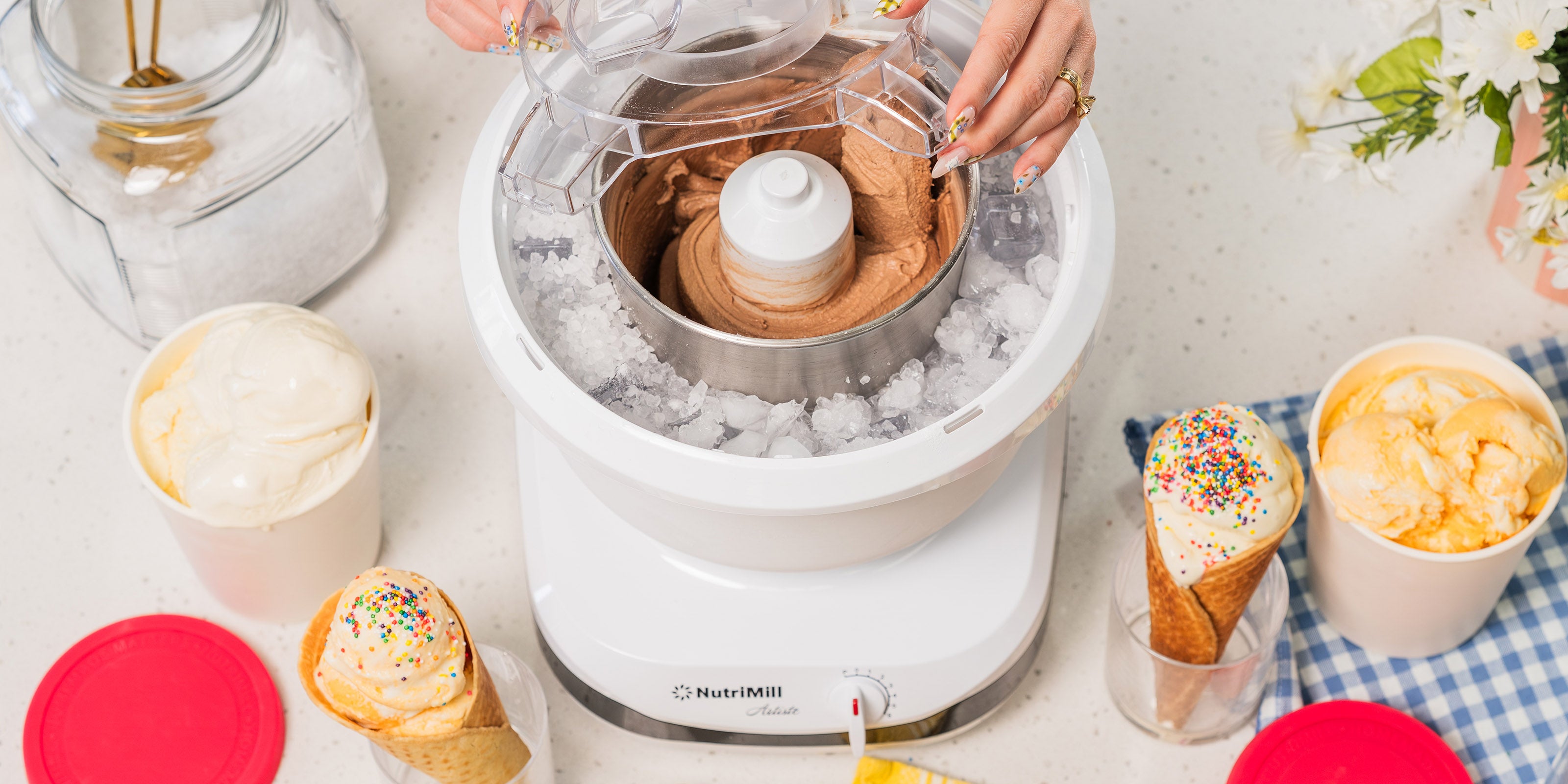 Lifestyle photo of hands using a white ice cream maker with ice cream cones and dishes nearby